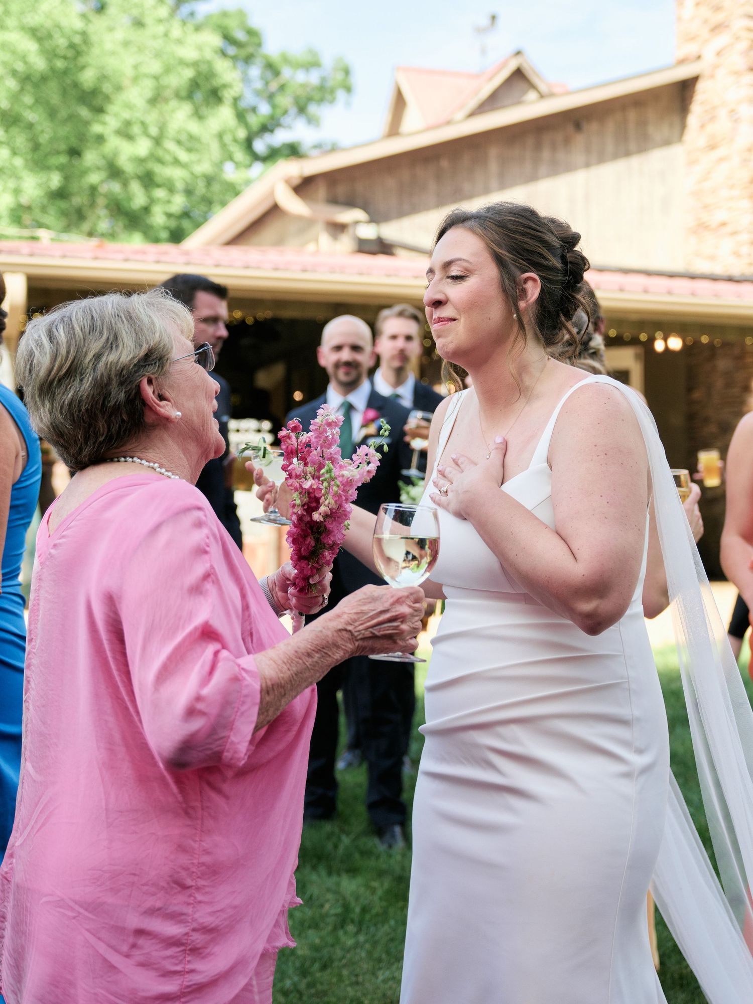 Two people share an animated conversation at an outdoor wedding reception with guests visible in the background.