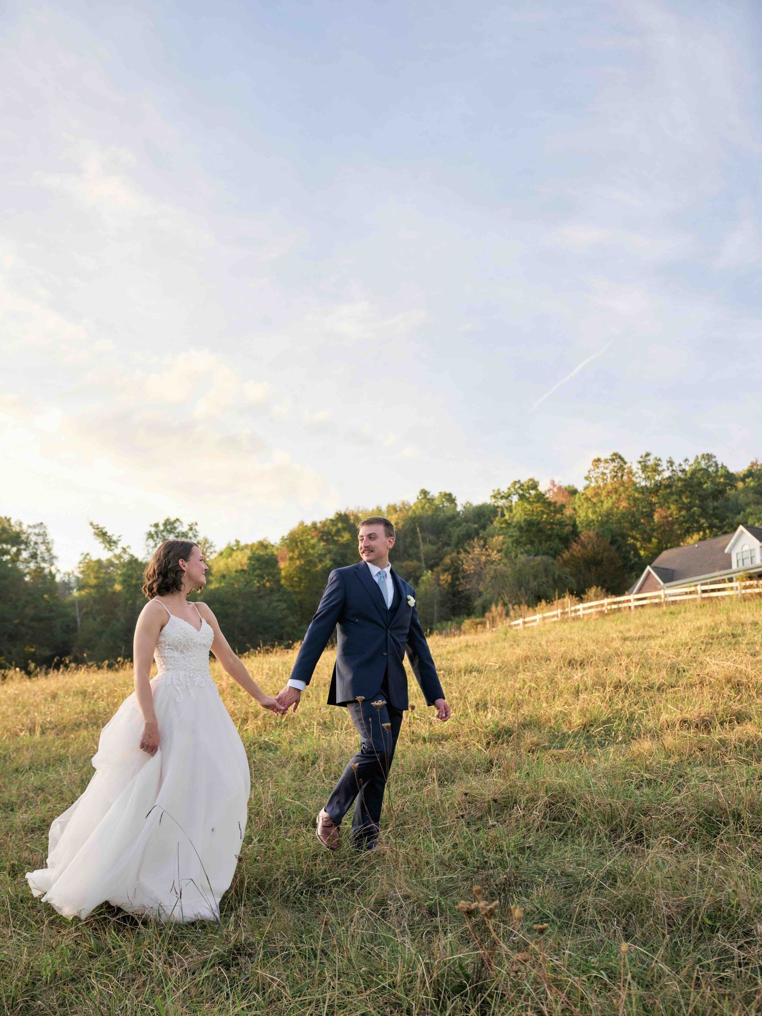 Bride and groom holding hands walking through golden field at sunset with rustic barn and wooded hillside backdrop.