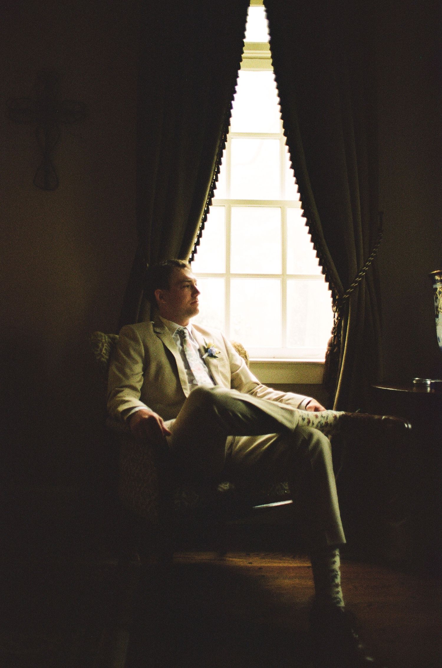 Man in light suit sitting thoughtfully in armchair by draped window in dimly lit elegant room.