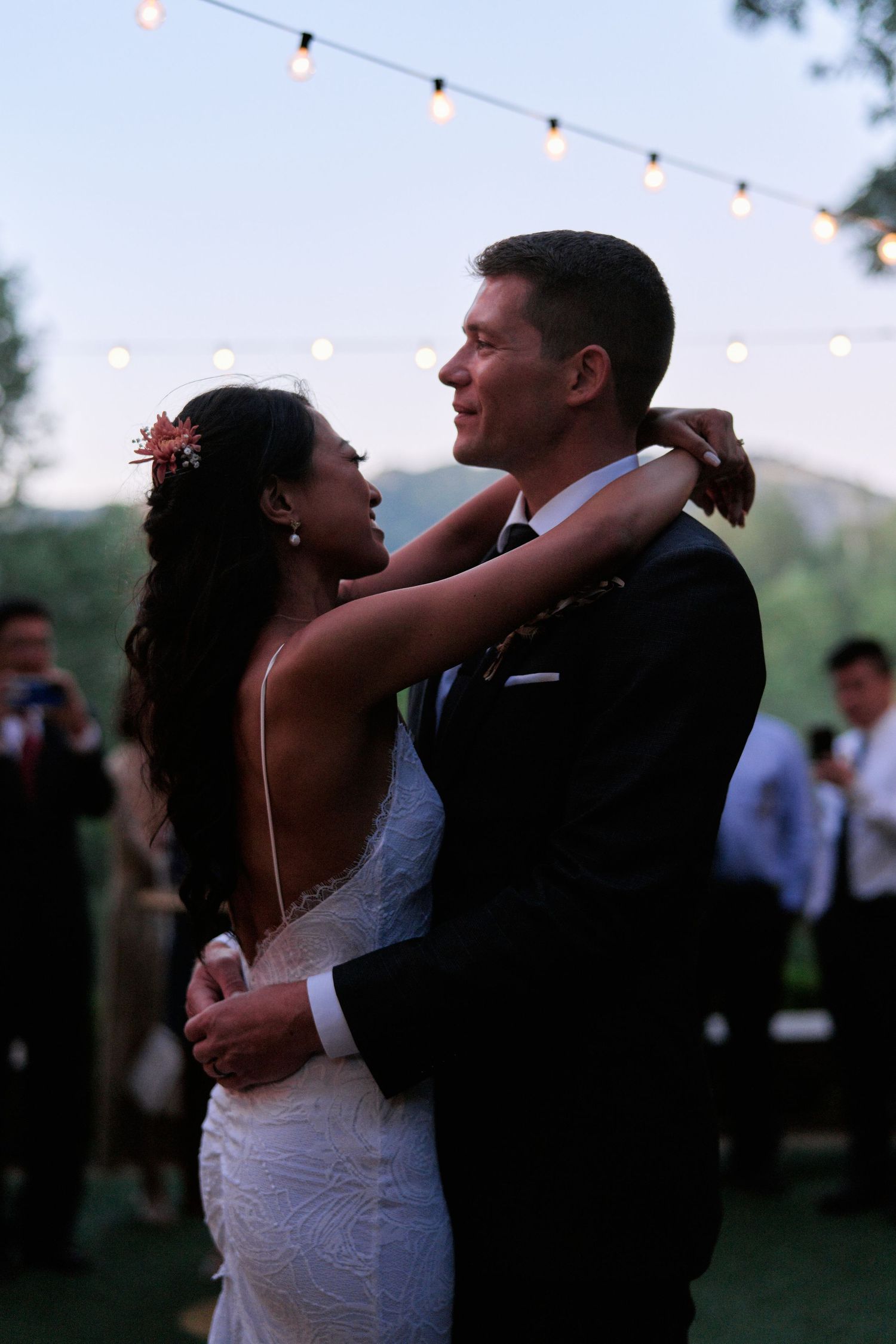 A couple shares their first dance outdoors under twinkling lights at twilight during their wedding reception.