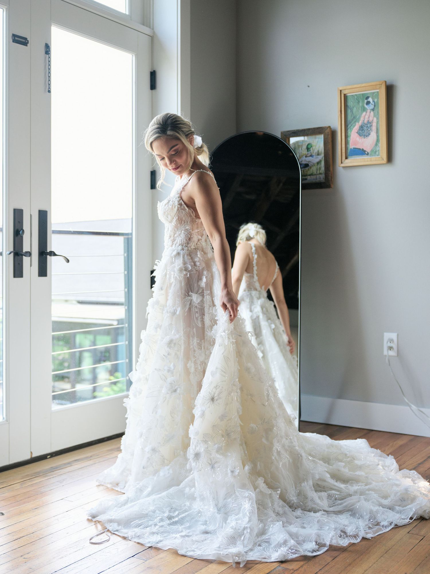 Two matching lace wedding dresses in white with long trains shown beside French doors in a sunlit room.