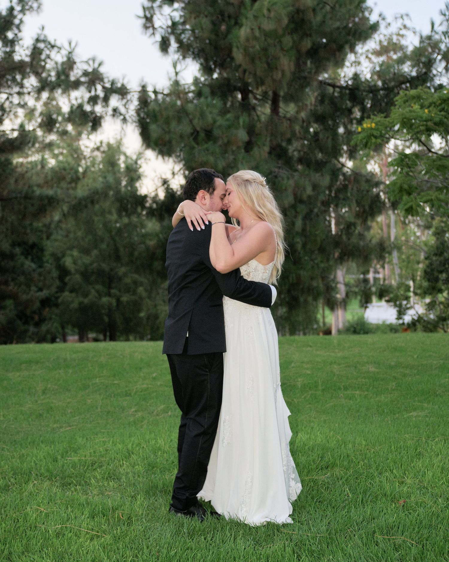 A tender wedding moment shared between newlyweds on a grassy lawn with trees in the background.