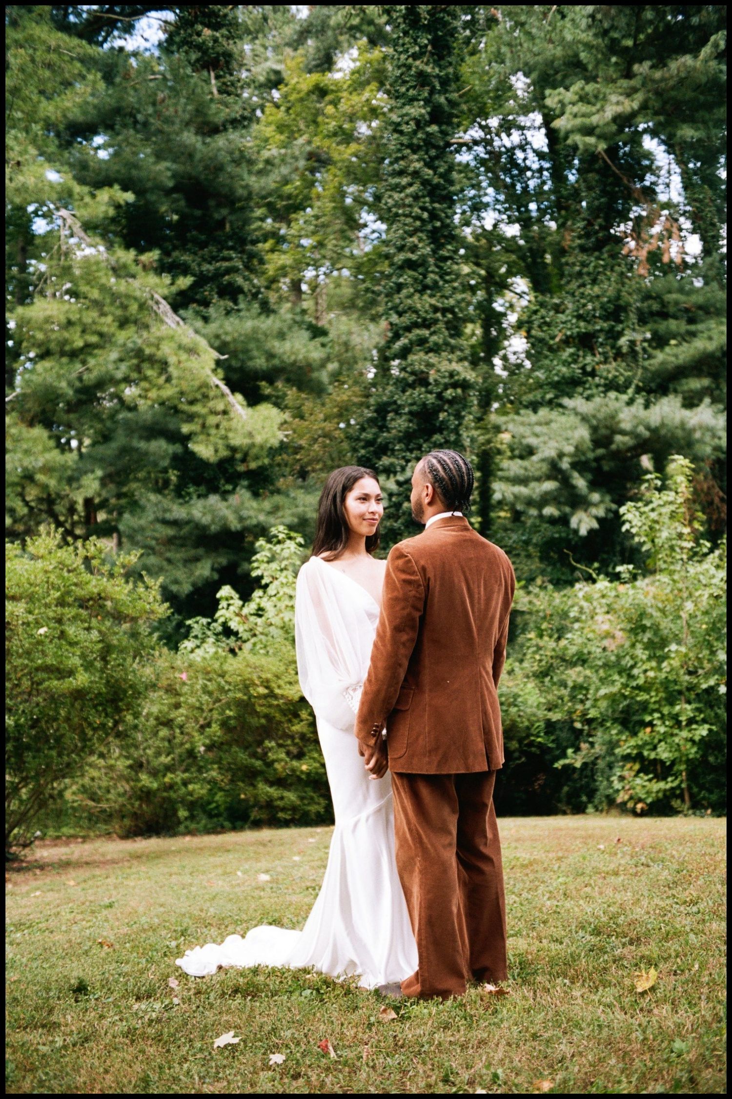 Bride and groom in brown suit share intimate moment in lush garden setting.