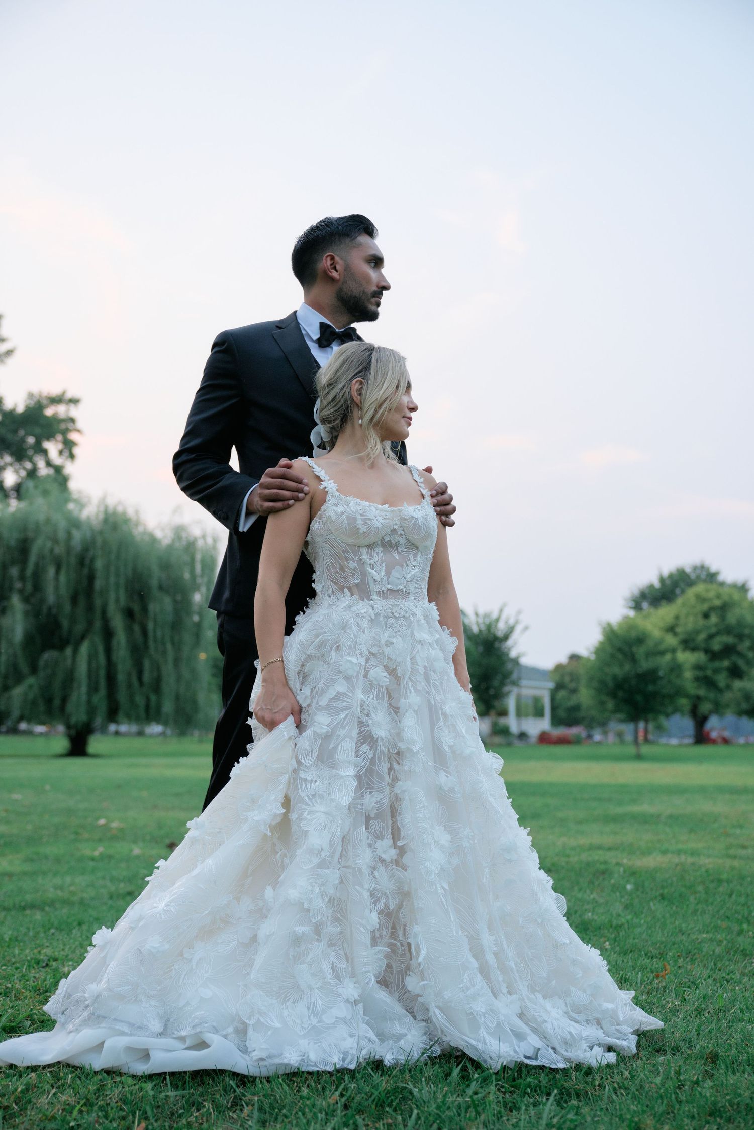 A wedding couple stands together on a lush lawn with willow trees in the background at sunset.