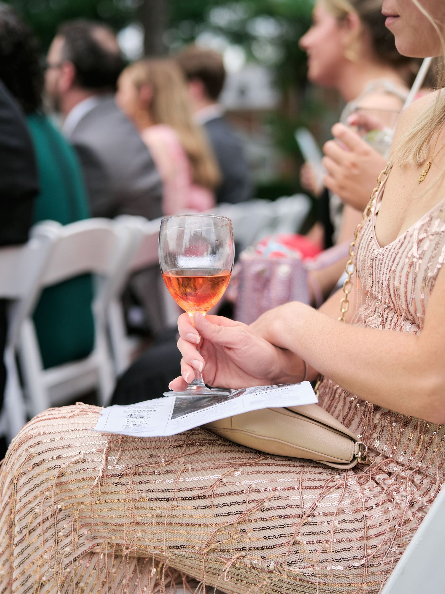 Guest holding a glass of rosé wine at an outdoor wedding reception while seated in a sequined dress.