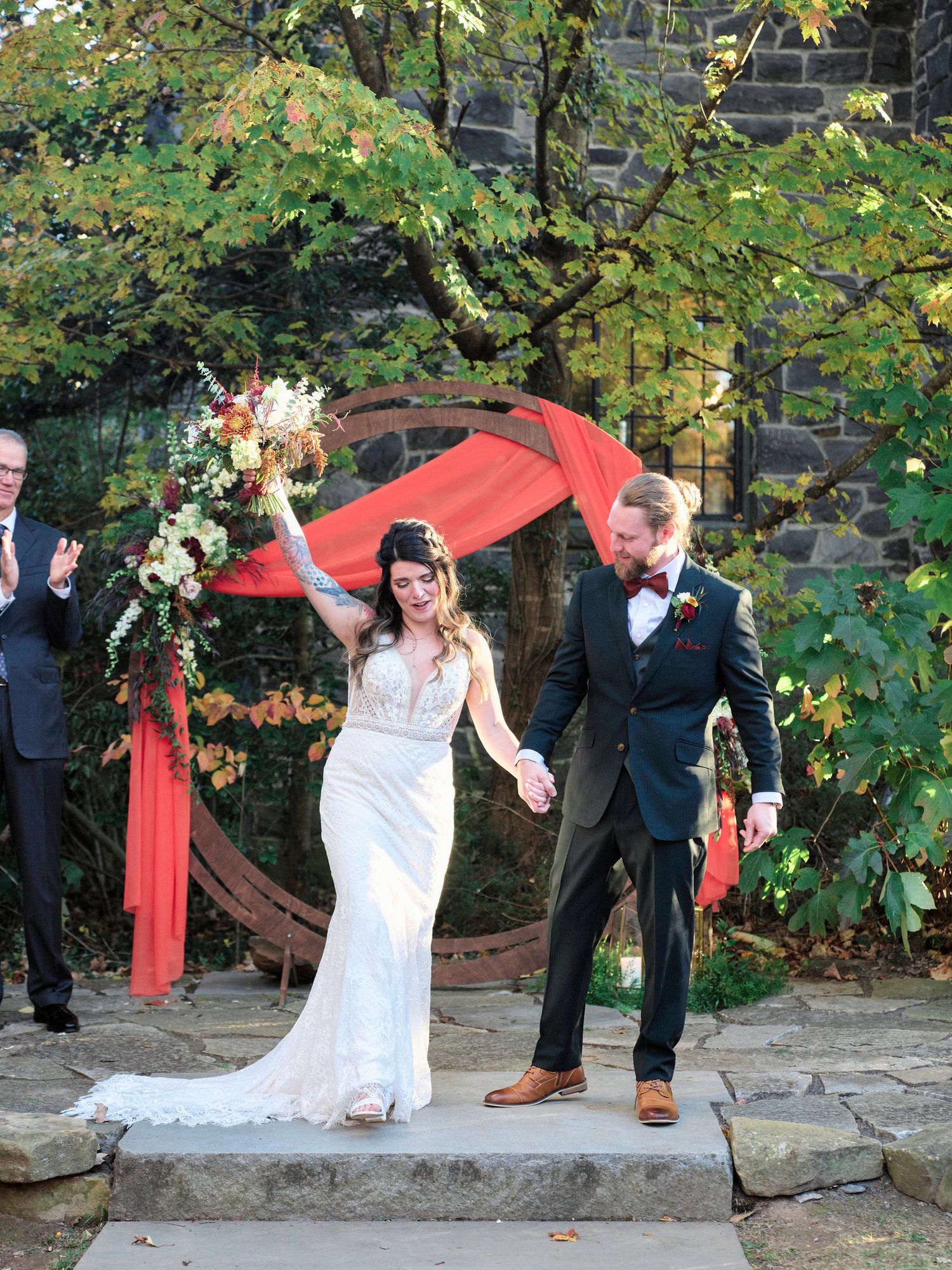 A couple celebrates their wedding exit under a red-draped archway surrounded by autumn foliage.