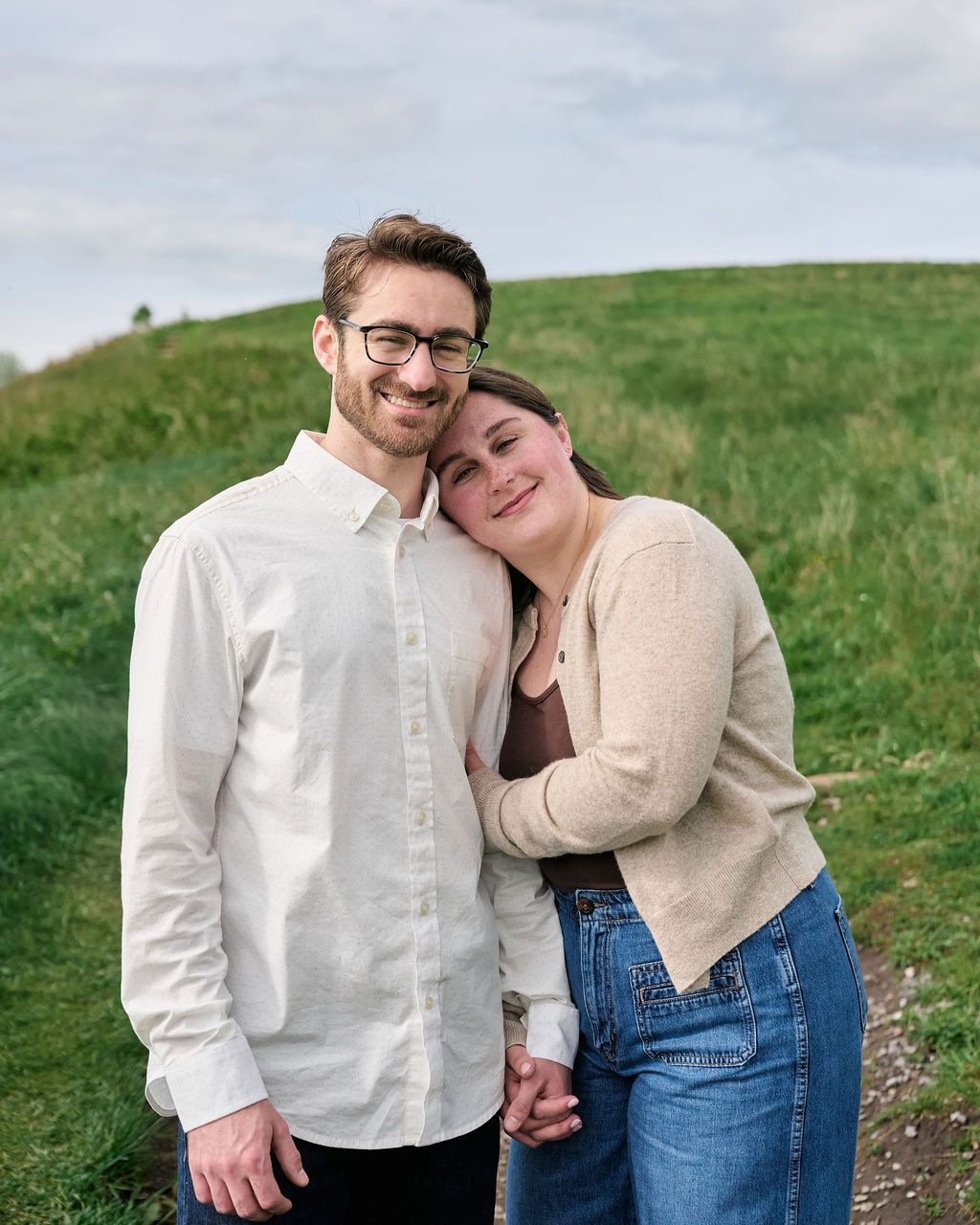 A playful couple in casual attire share joyful moments against a grassy hillside backdrop.