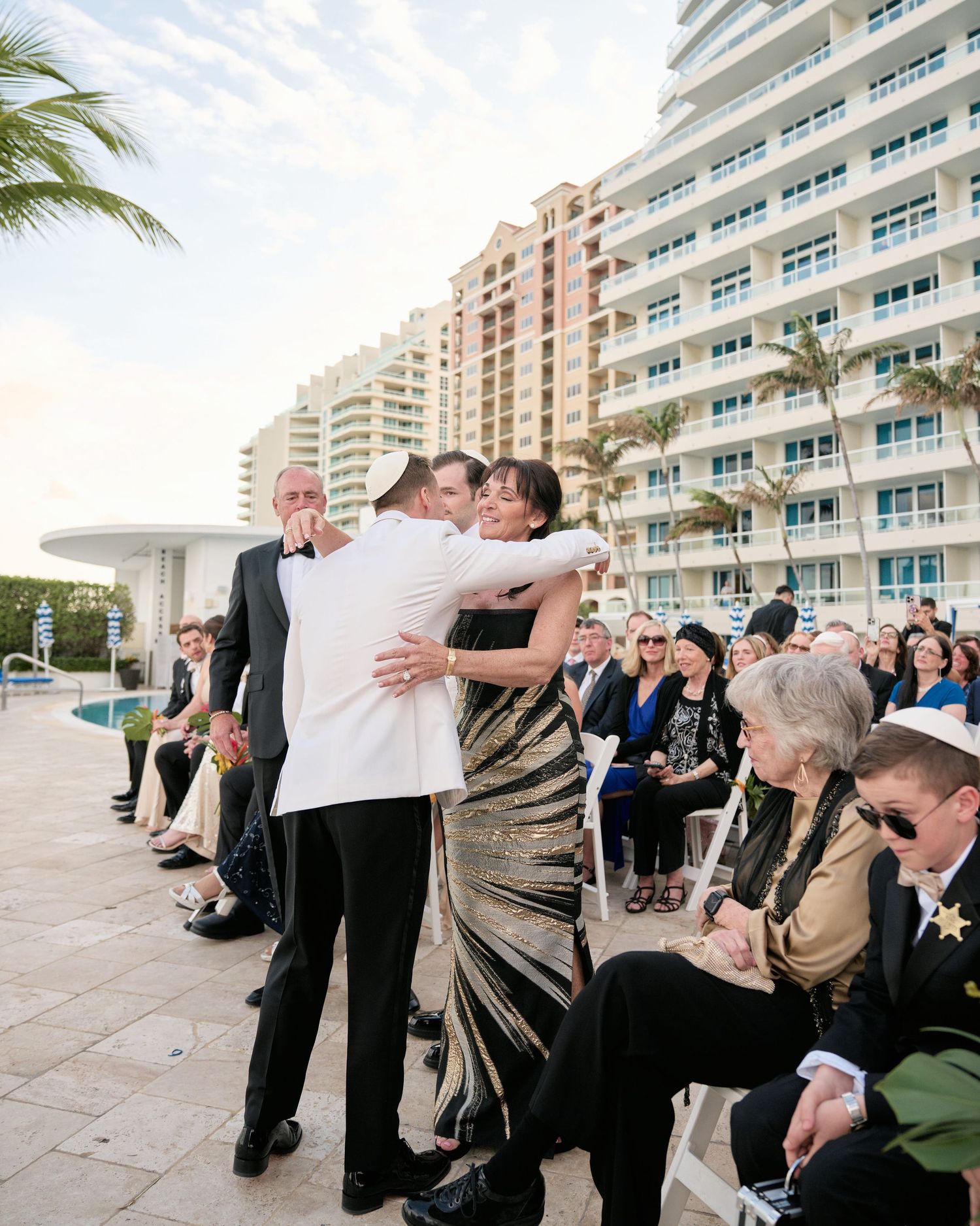 Outdoor wedding ceremony with guests seated on white chairs beside tall beige apartment buildings and palm trees.