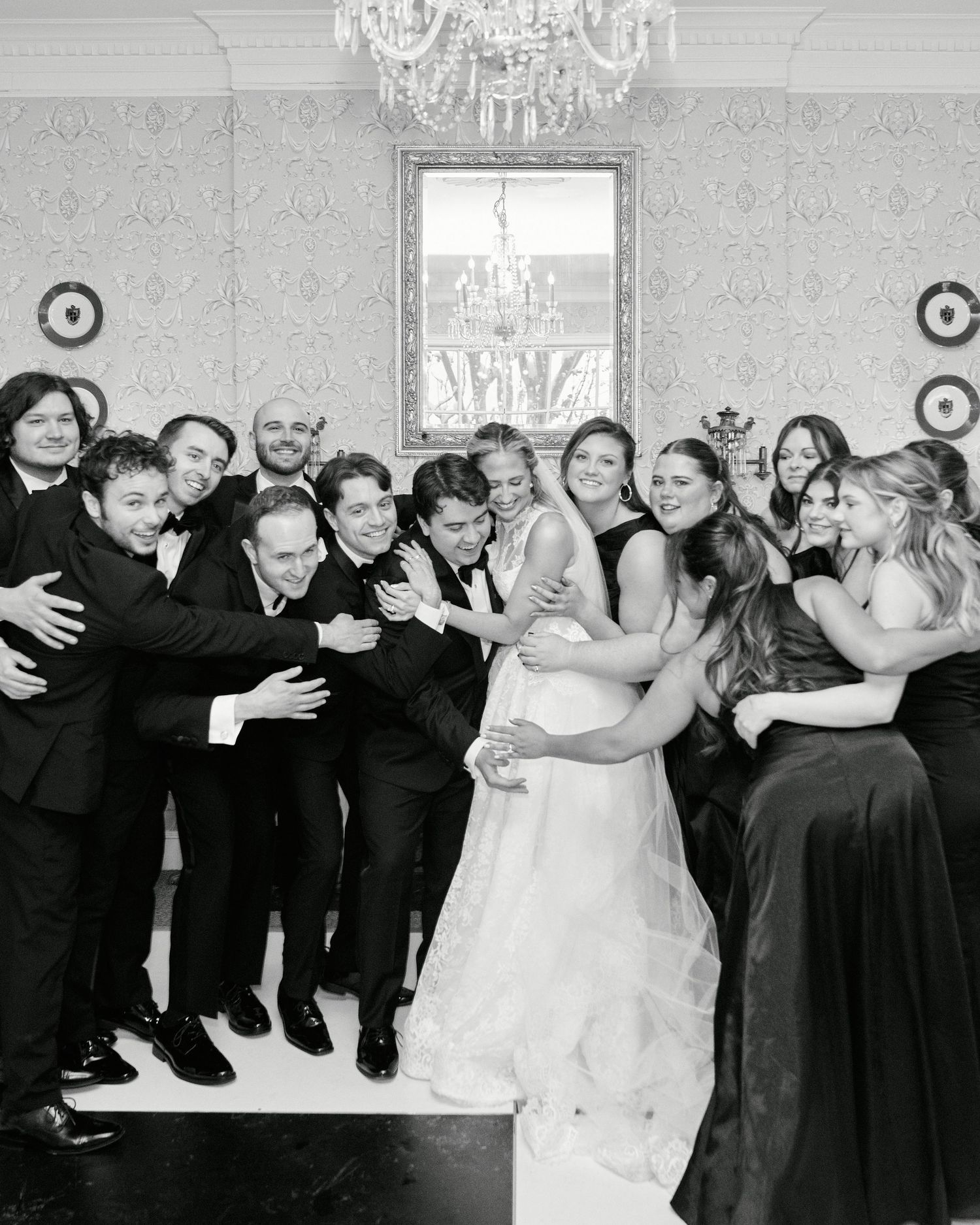 Black and white wedding party photo with bride, groom, and guests hugging in elegant chandelier ballroom.