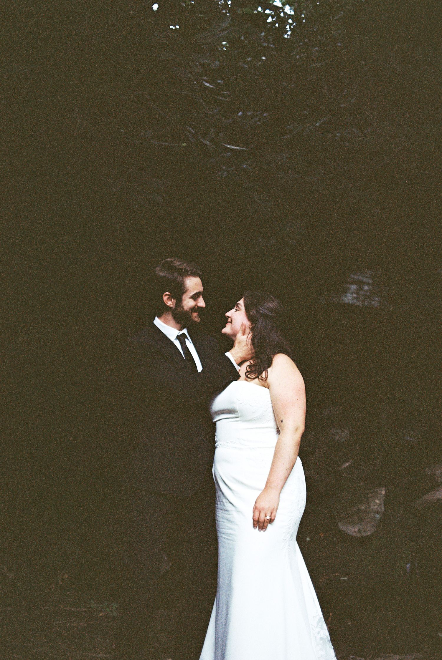 A laughing bride in white gown and groom in black suit share a joyful moment outdoors in natural light.