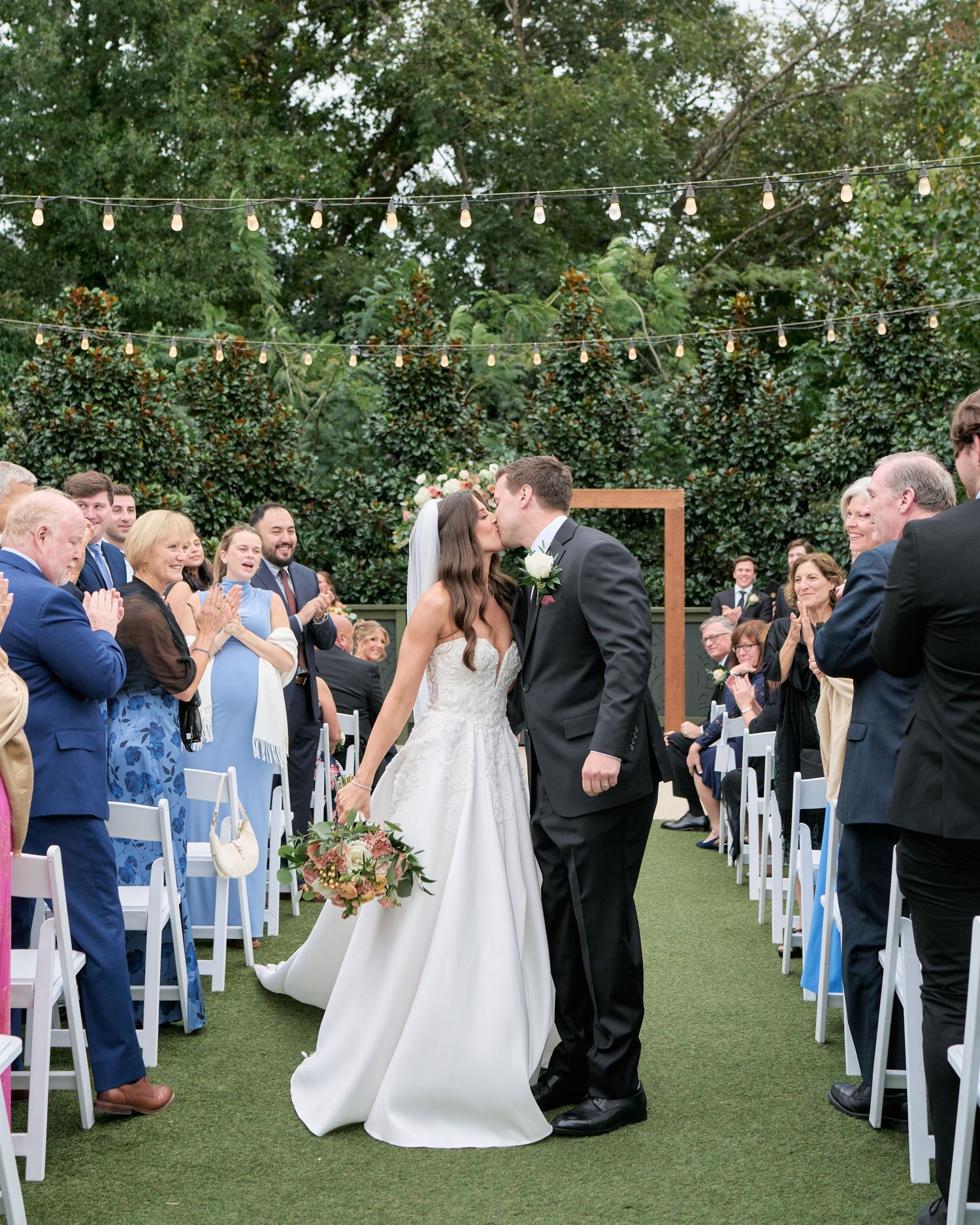 A romantic outdoor wedding ceremony with string lights overhead and white chairs lining the aisle on a green lawn.