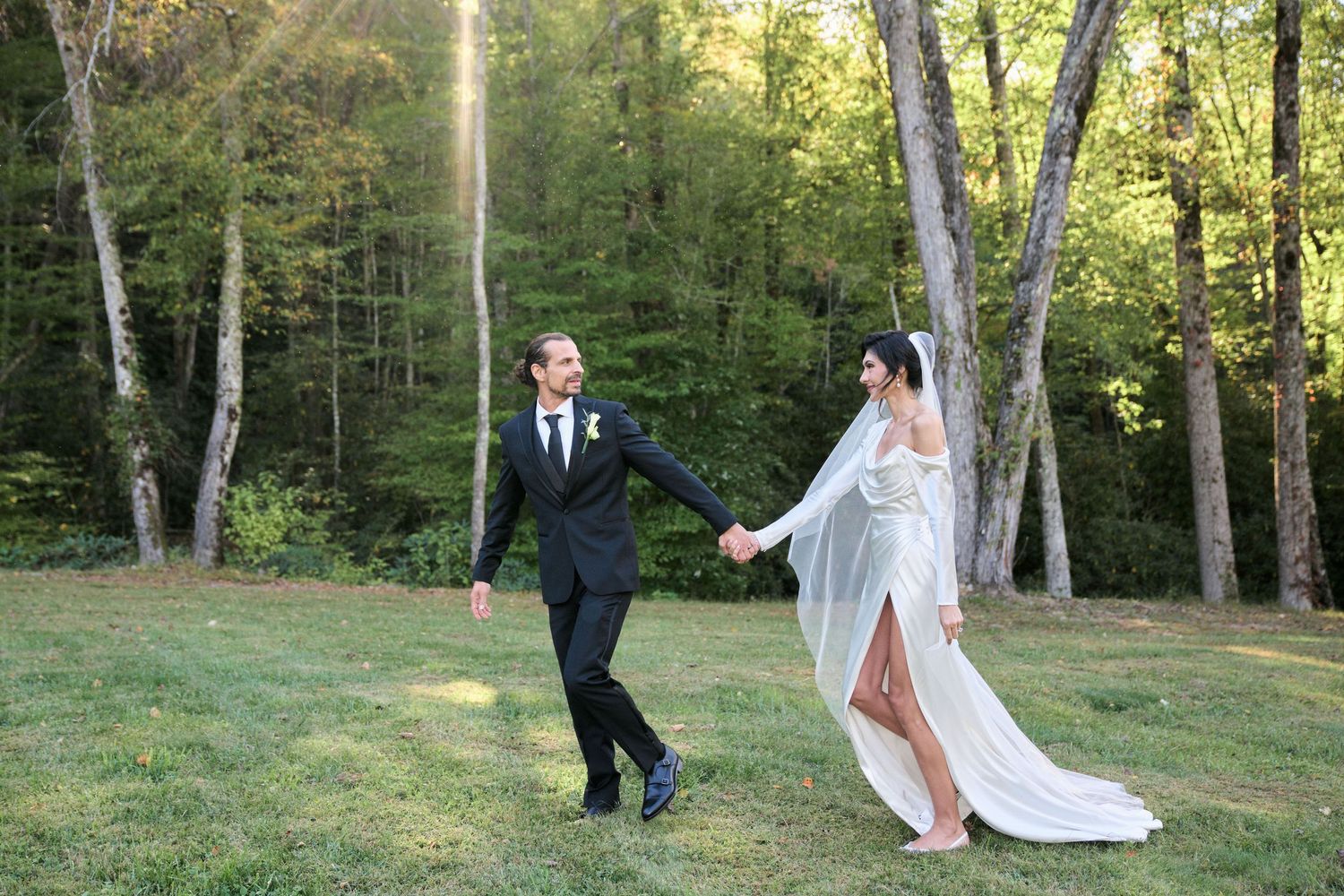 A couple walks through a scenic wooded area in wedding attire as their dress trails behind them on the grass.