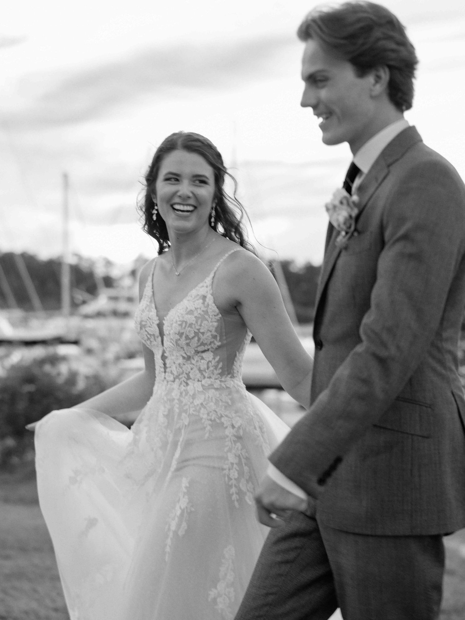 Black and white photo of a laughing bride in lace gown holding hands with groom in gray suit at waterfront wedding.