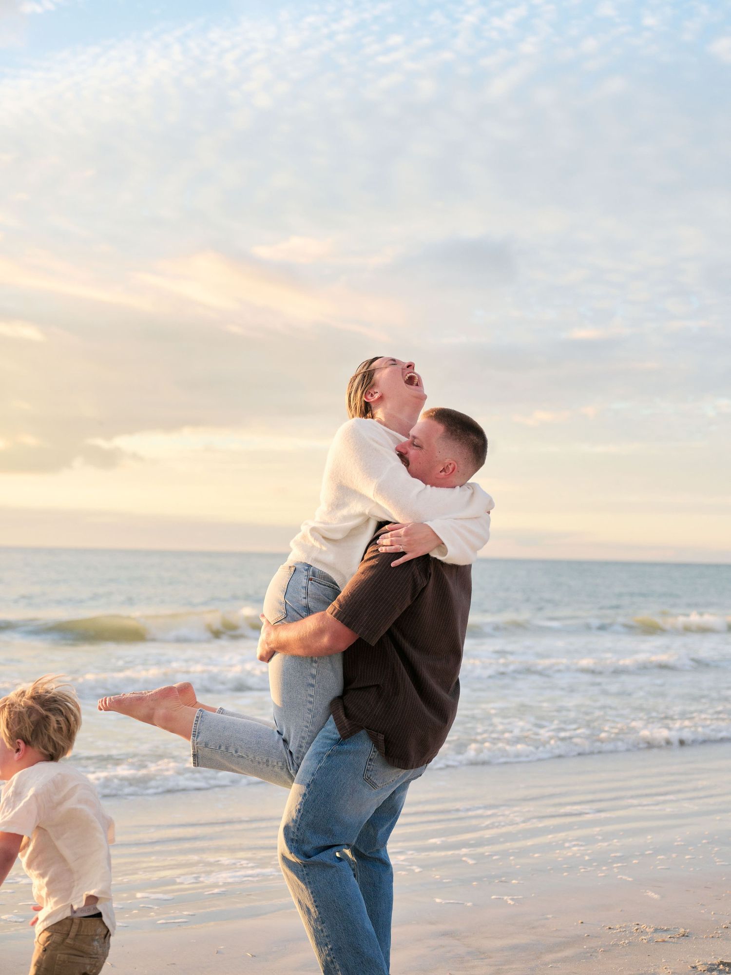 A joyful couple embraces on the beach at sunset while a child plays nearby.