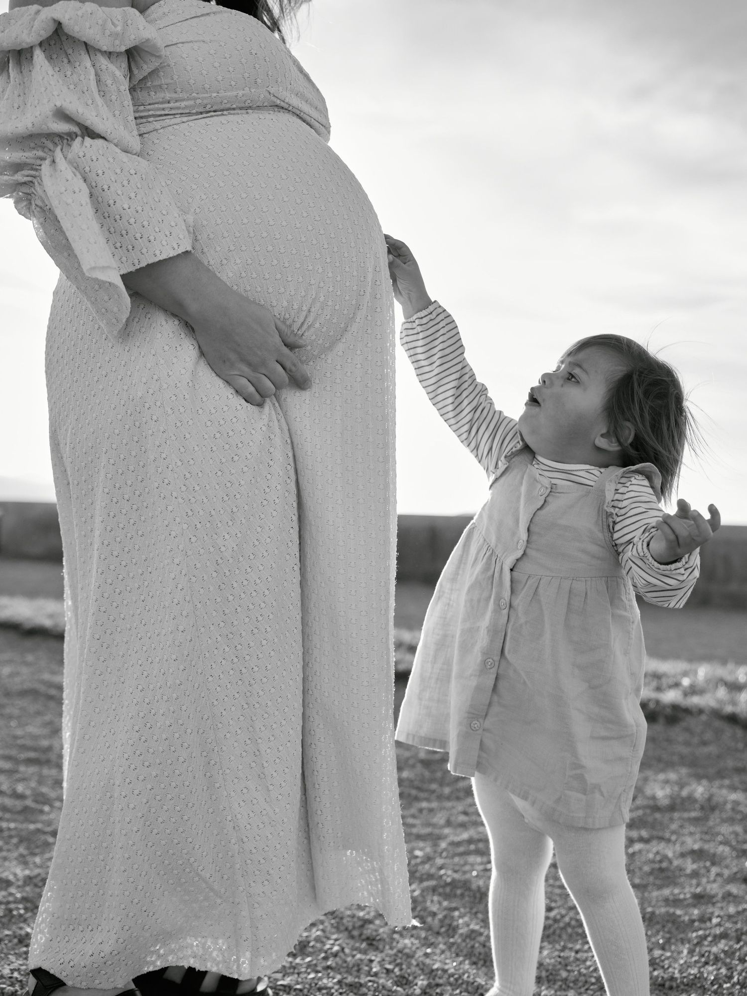 Black and white maternity photo of pregnant woman in white dress with toddler girl reaching up outdoors.