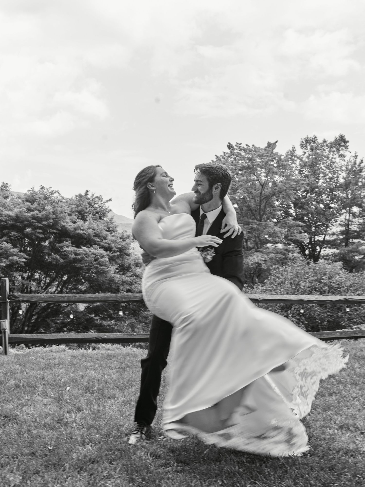 Black and white photo of a groom lifting his bride in a wedding dress outdoors, both laughing joyfully.