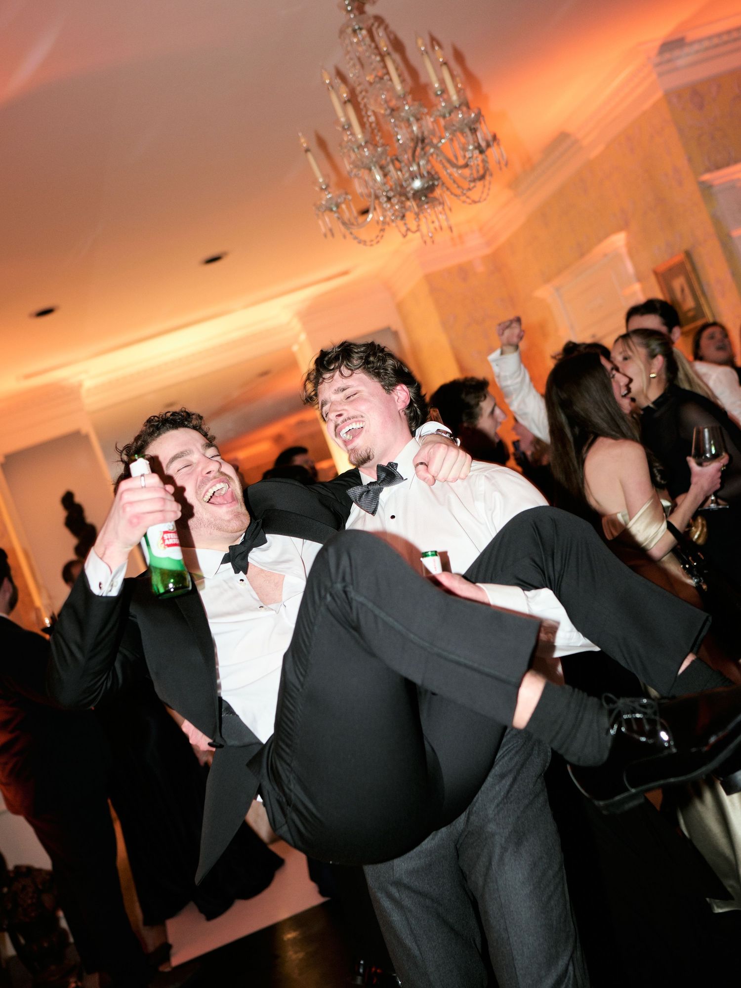 Two men in tuxedos celebrate at a formal event, one carrying the other while holding drinks on a dance floor.