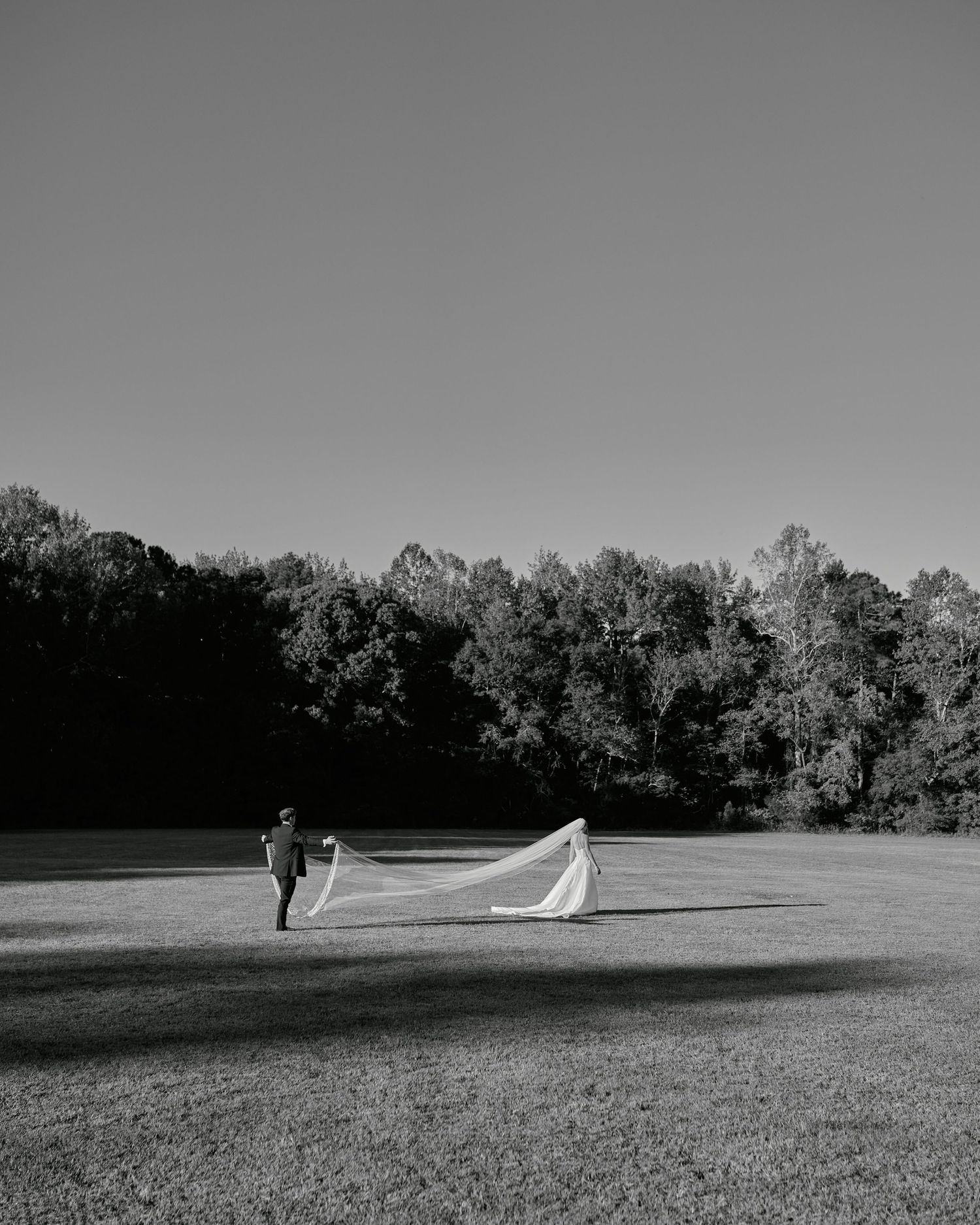A black and white photo of a couple walking across a field with a flowing wedding dress train trailing behind them.