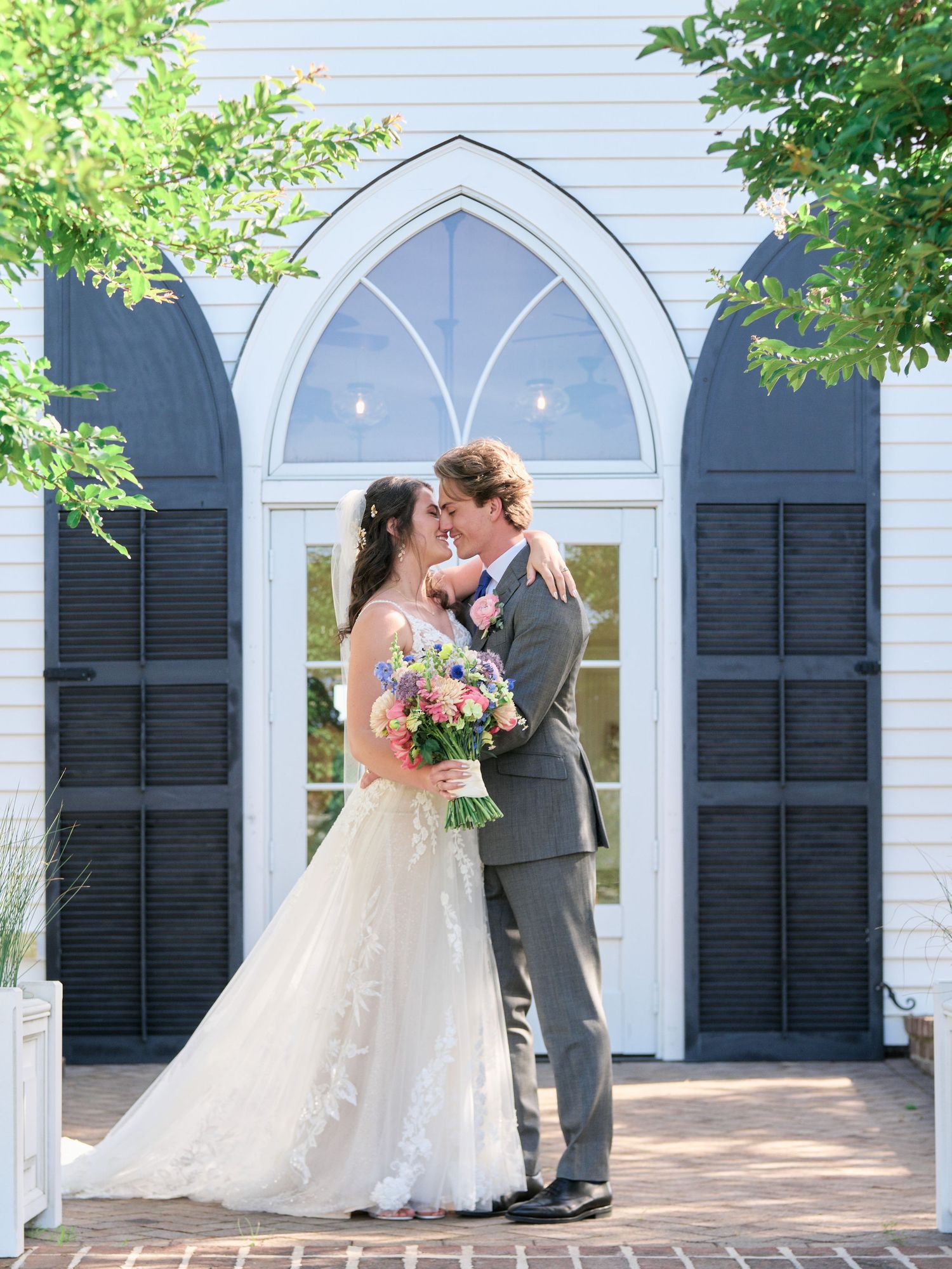 Bride and groom share a romantic embrace in front of a white chapel with gothic arched windows on their wedding day.