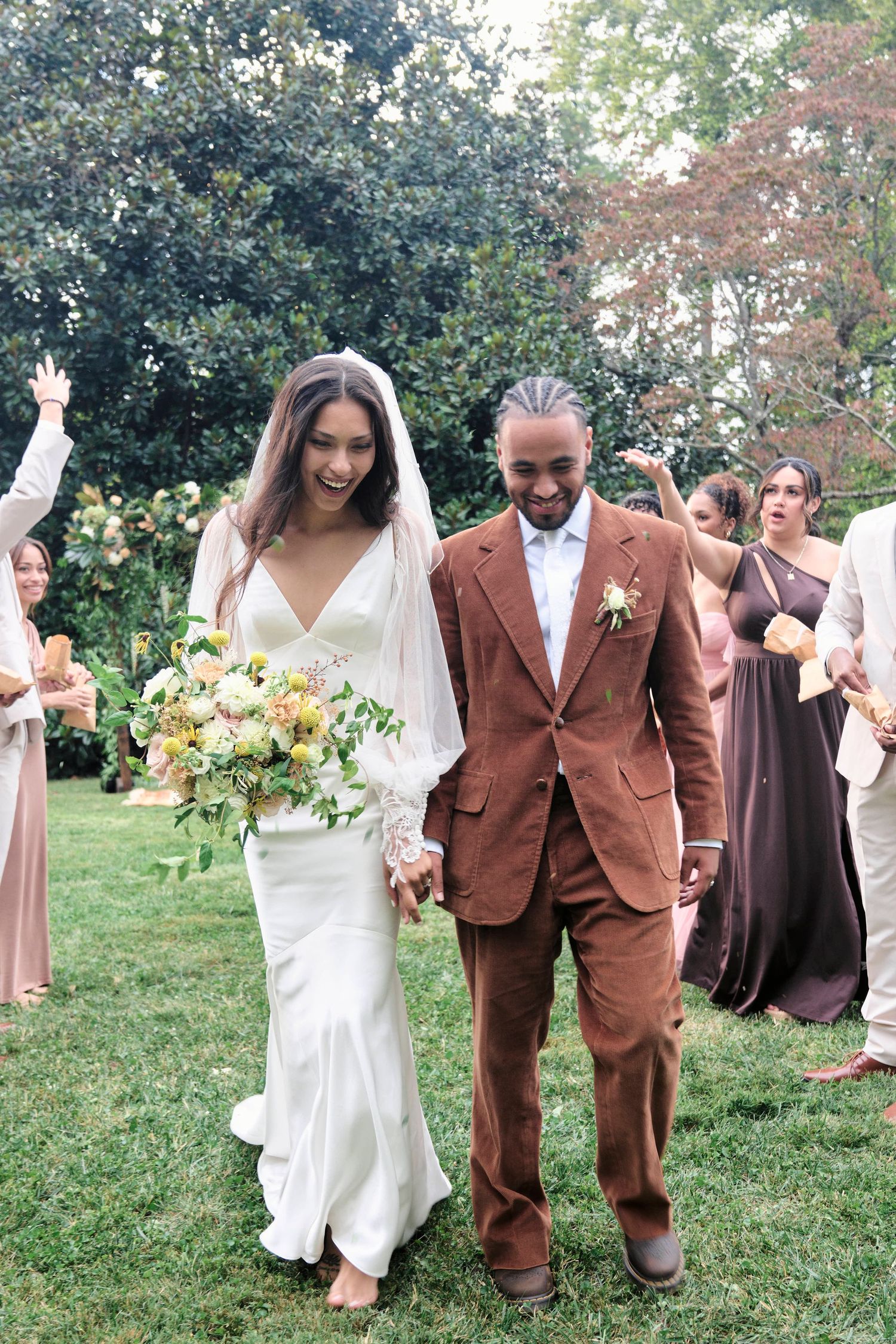A wedding couple walks across a lawn while guests in formal attire celebrate in the background.