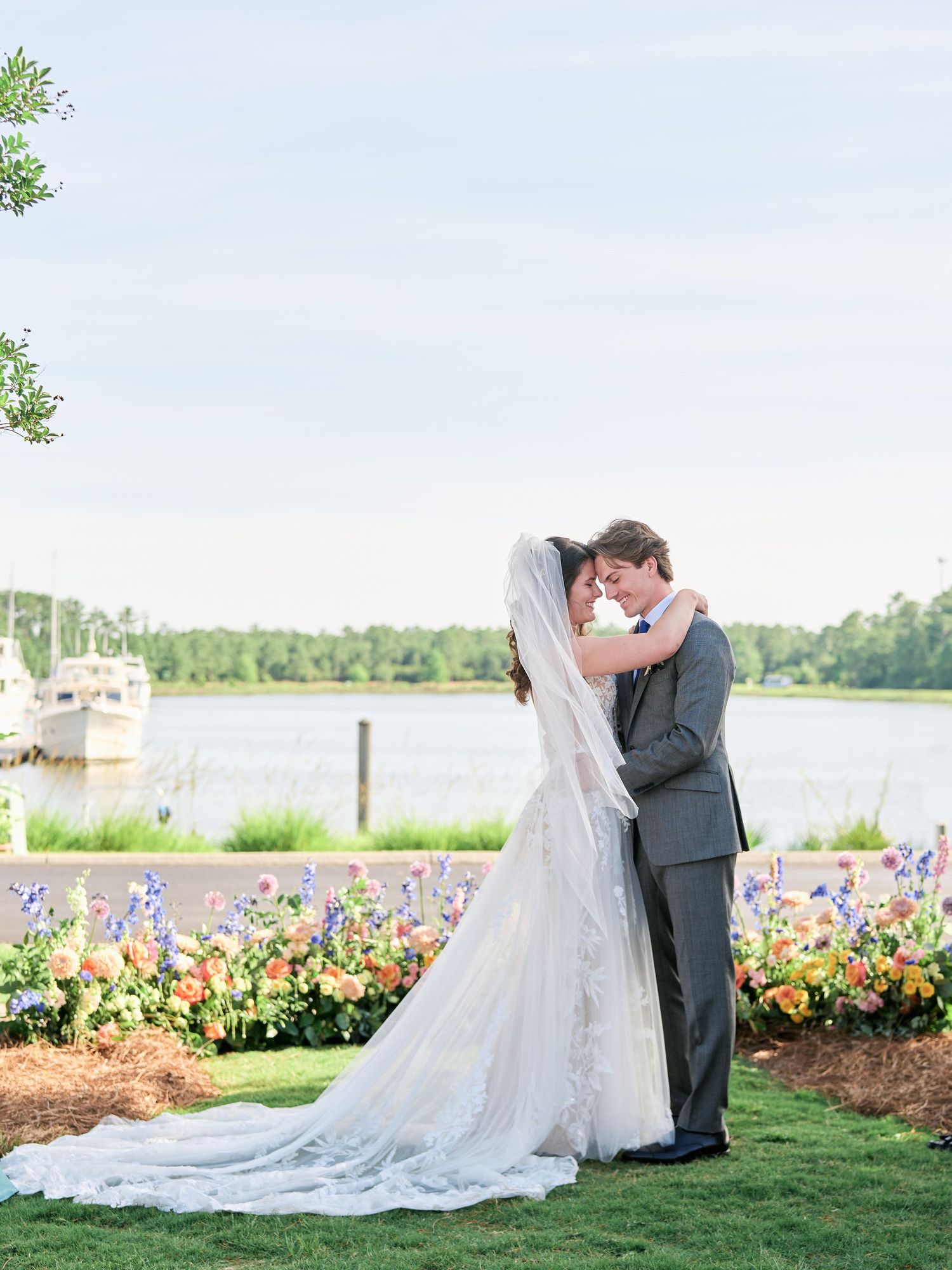 Newlyweds share romantic moment by lakeside garden with colorful flower beds and water view on sunny day.