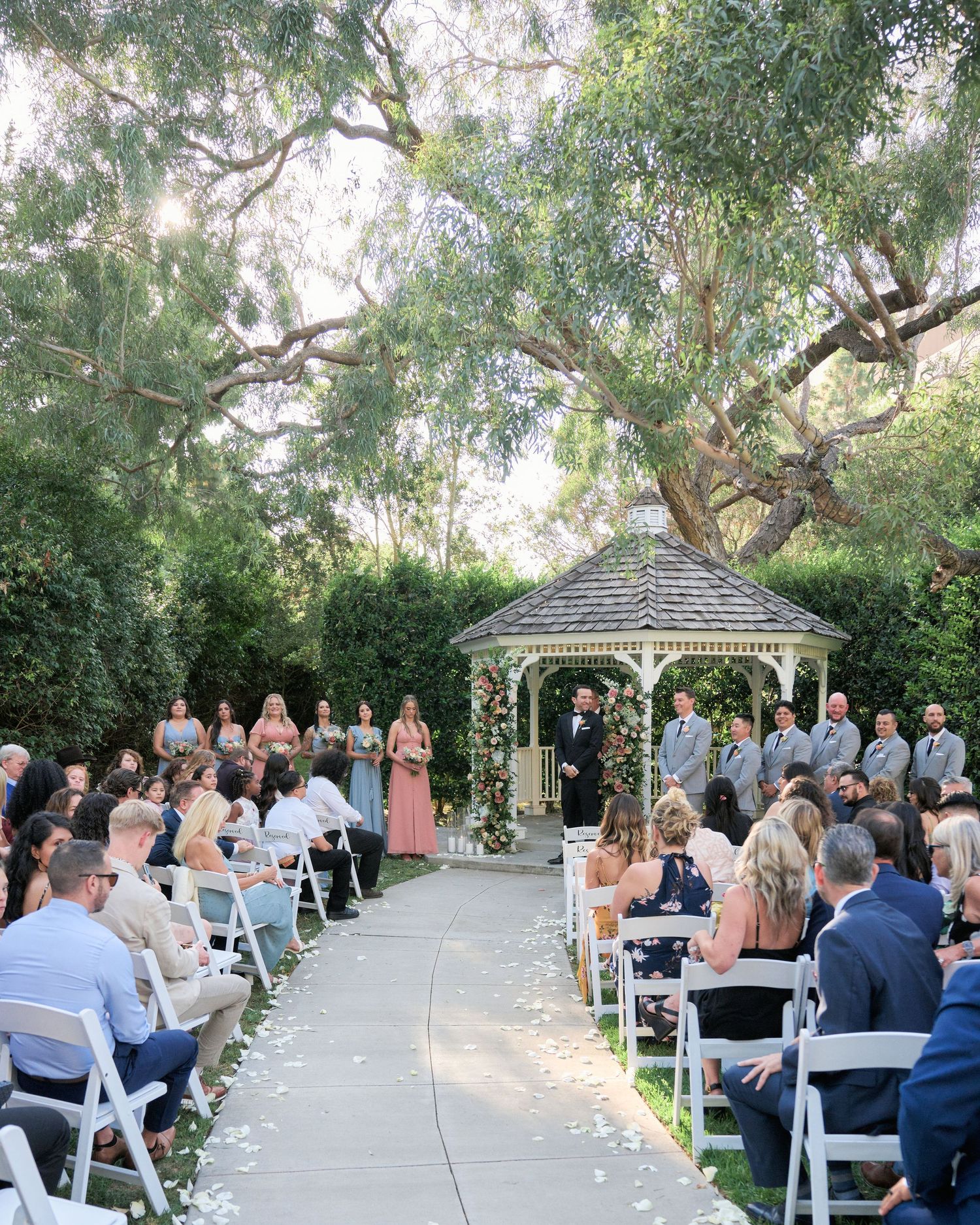 Outdoor wedding ceremony at a garden gazebo with guests seated on white chairs beneath towering trees.