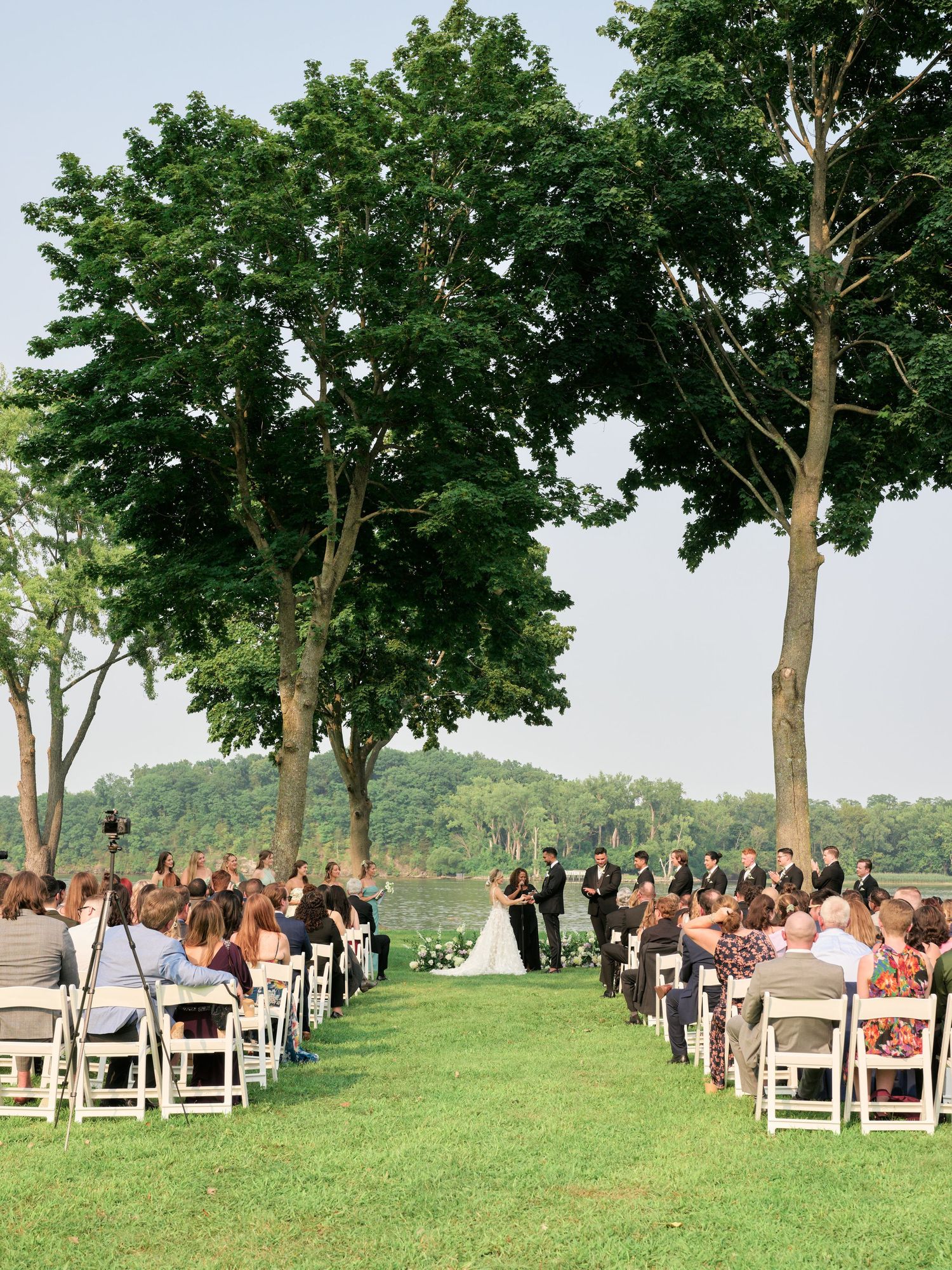 Outdoor wedding ceremony setup with white chairs arranged beneath tall trees on a grassy hilltop overlooking scenic views.
