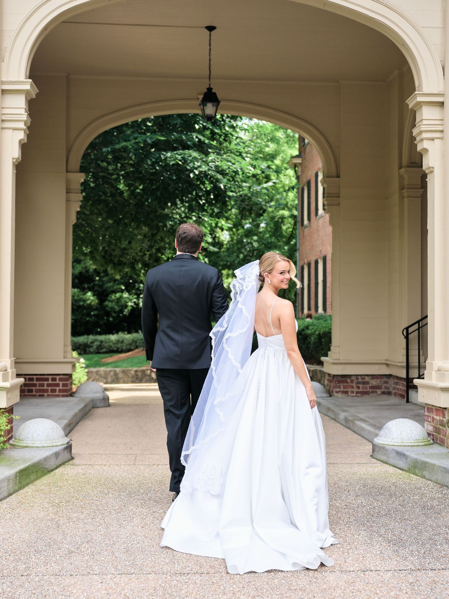 A bride in flowing white gown and groom walking away under an arched colonnade.
