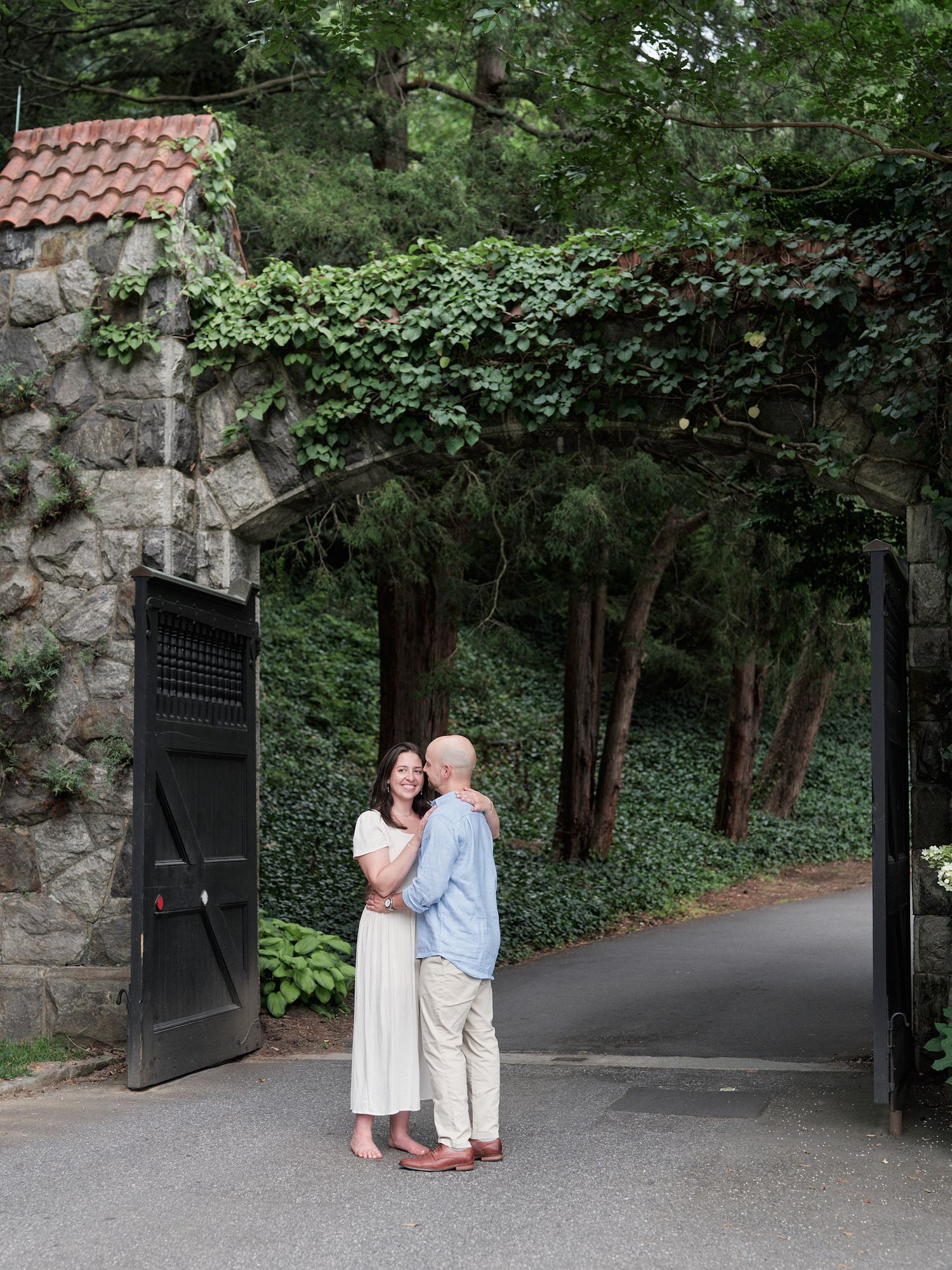 A couple poses in front of a stone gateway entrance with ivy growing on the walls.
