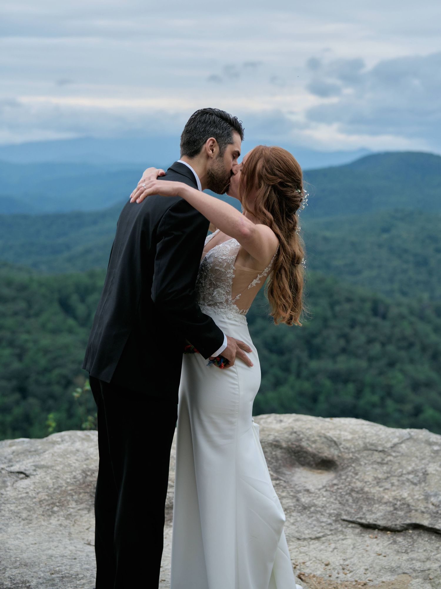 A couple shares a romantic kiss on a mountain overlook with sweeping forest views and cloudy skies in the background.