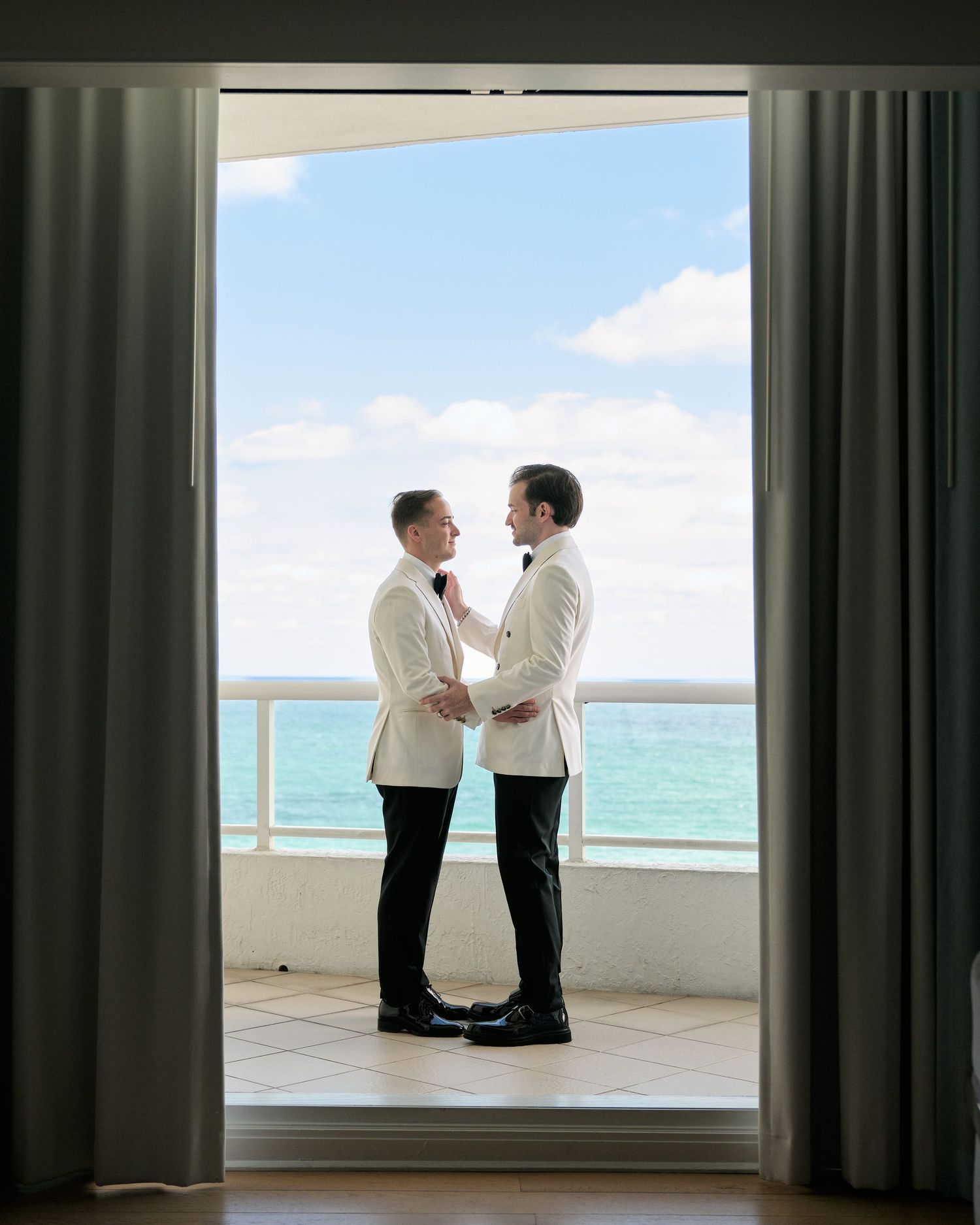 Two people in cream jackets share an intimate moment on a balcony framed by curtains.