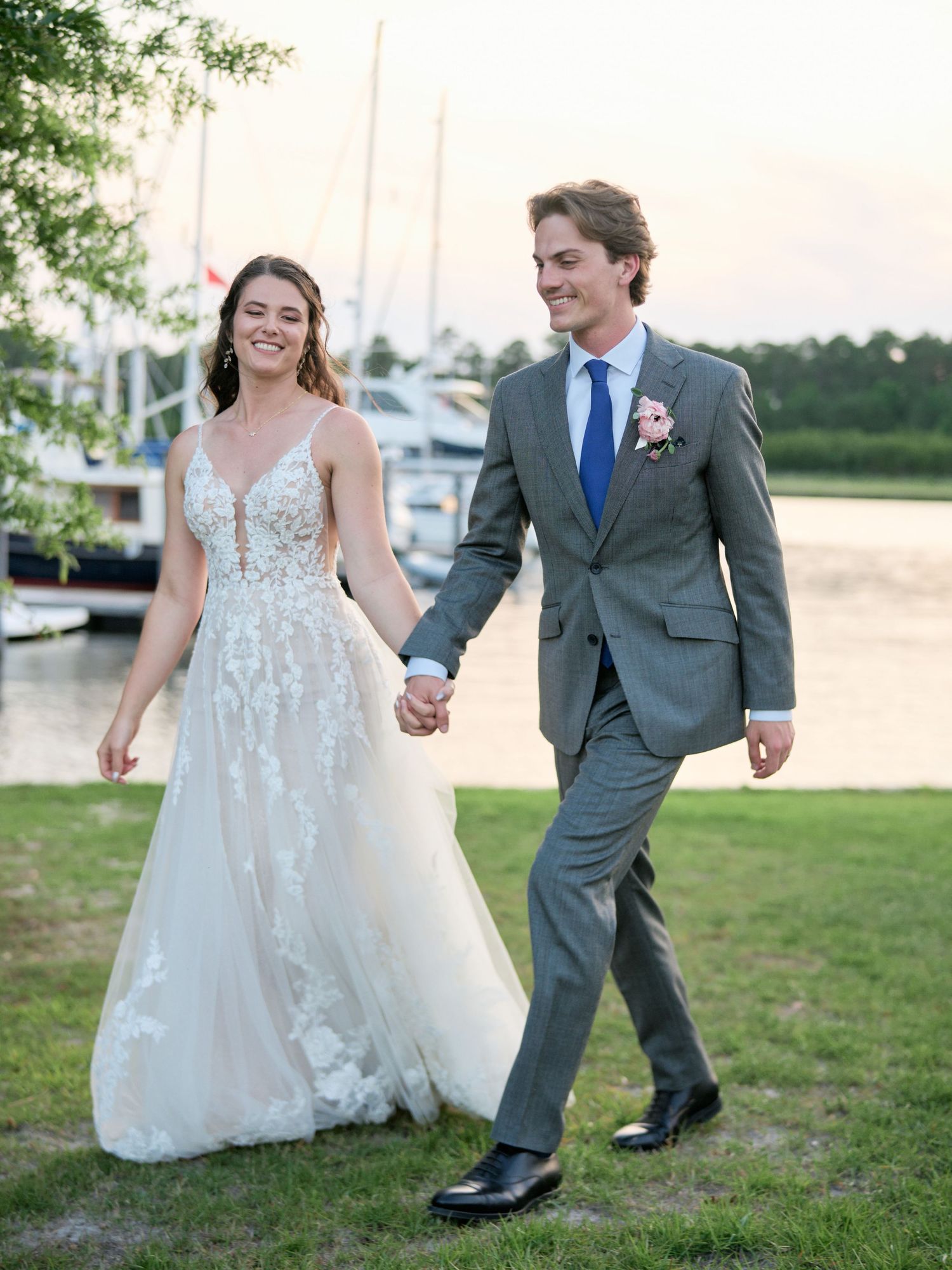 A newlywed couple walks hand in hand along a marina with boats and greenery in the background at sunset.