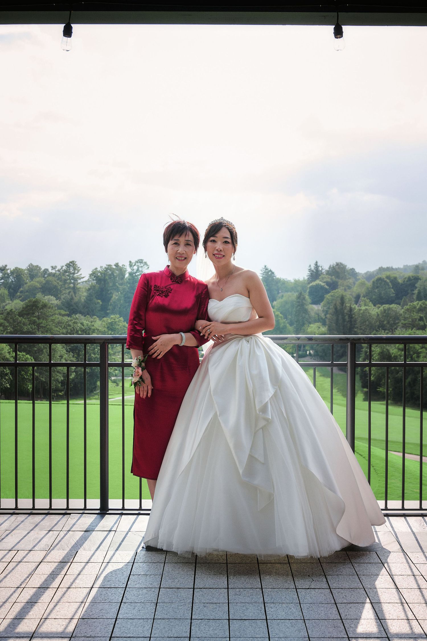 A bride in a ballgown and woman in red dress pose together on a balcony overlooking a golf course.