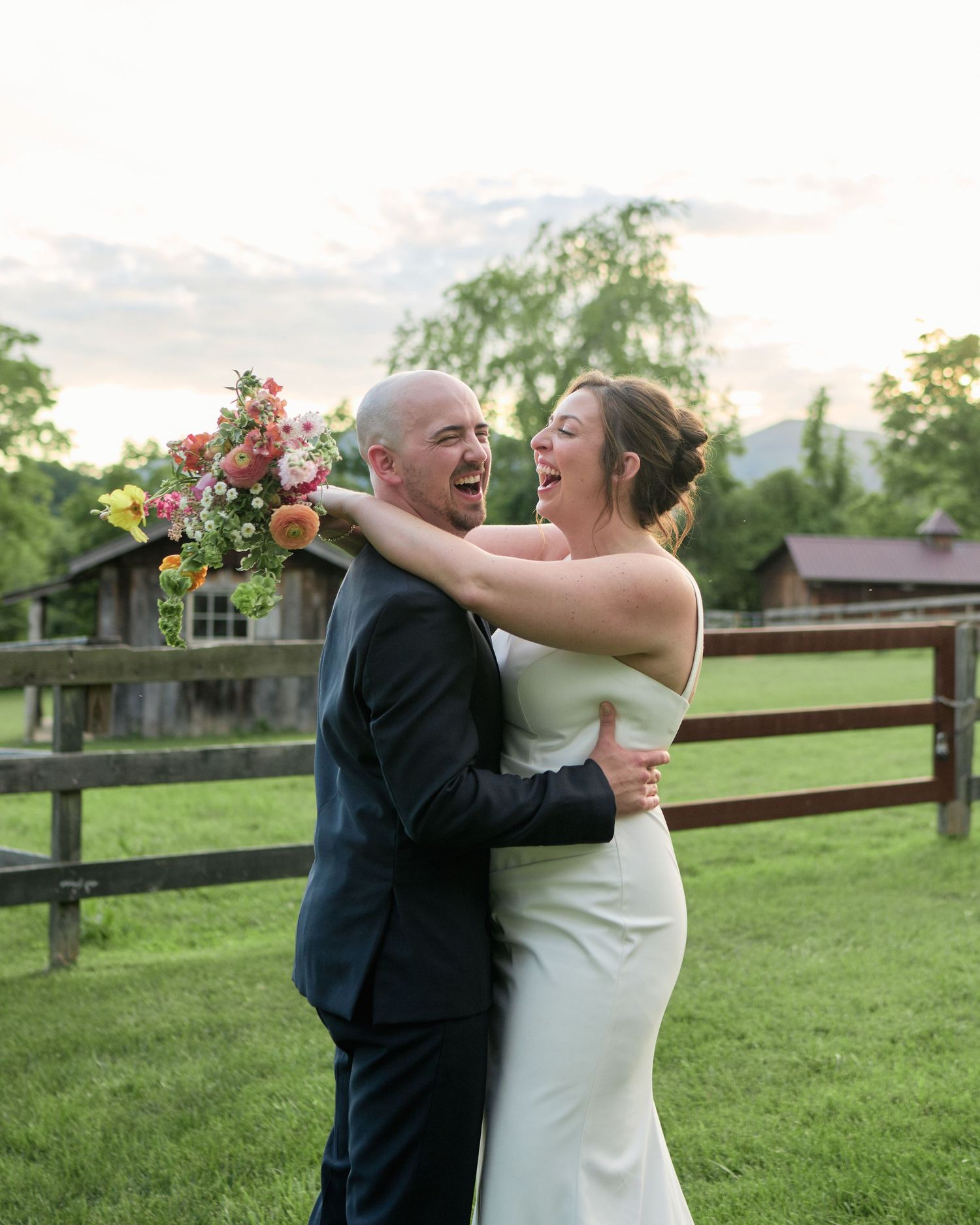 A joyful wedding couple embraces and laughs together in front of a rustic fence at sunset.