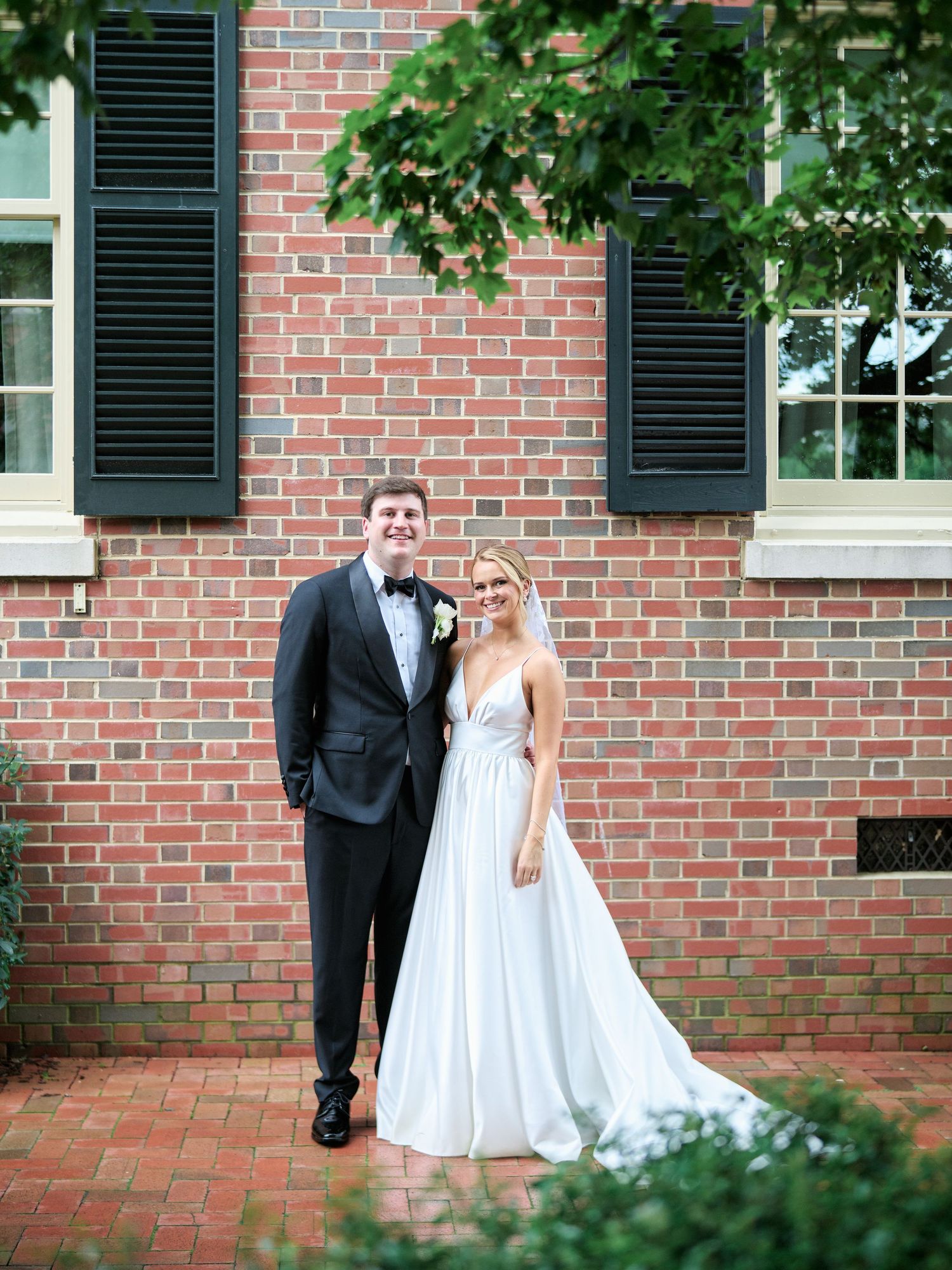 Elegant wedding couple poses in front of a brick building with black shutters.
