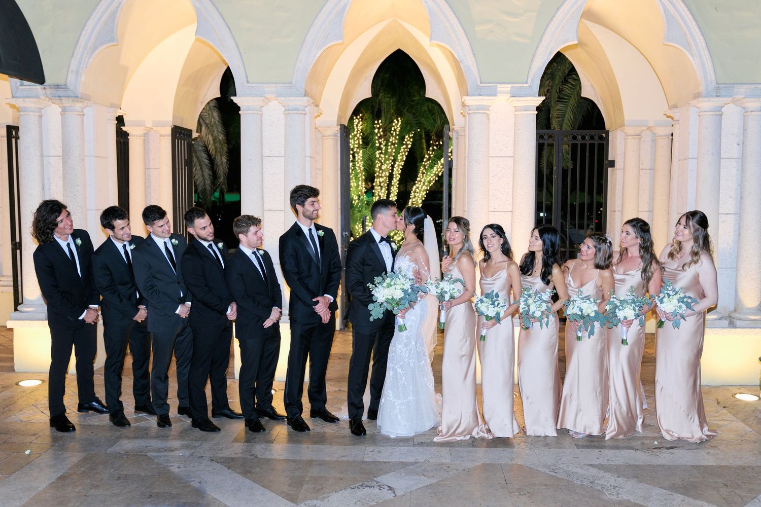 Wedding party poses under elegant arches with bridesmaids in blush and groomsmen in dark suits.