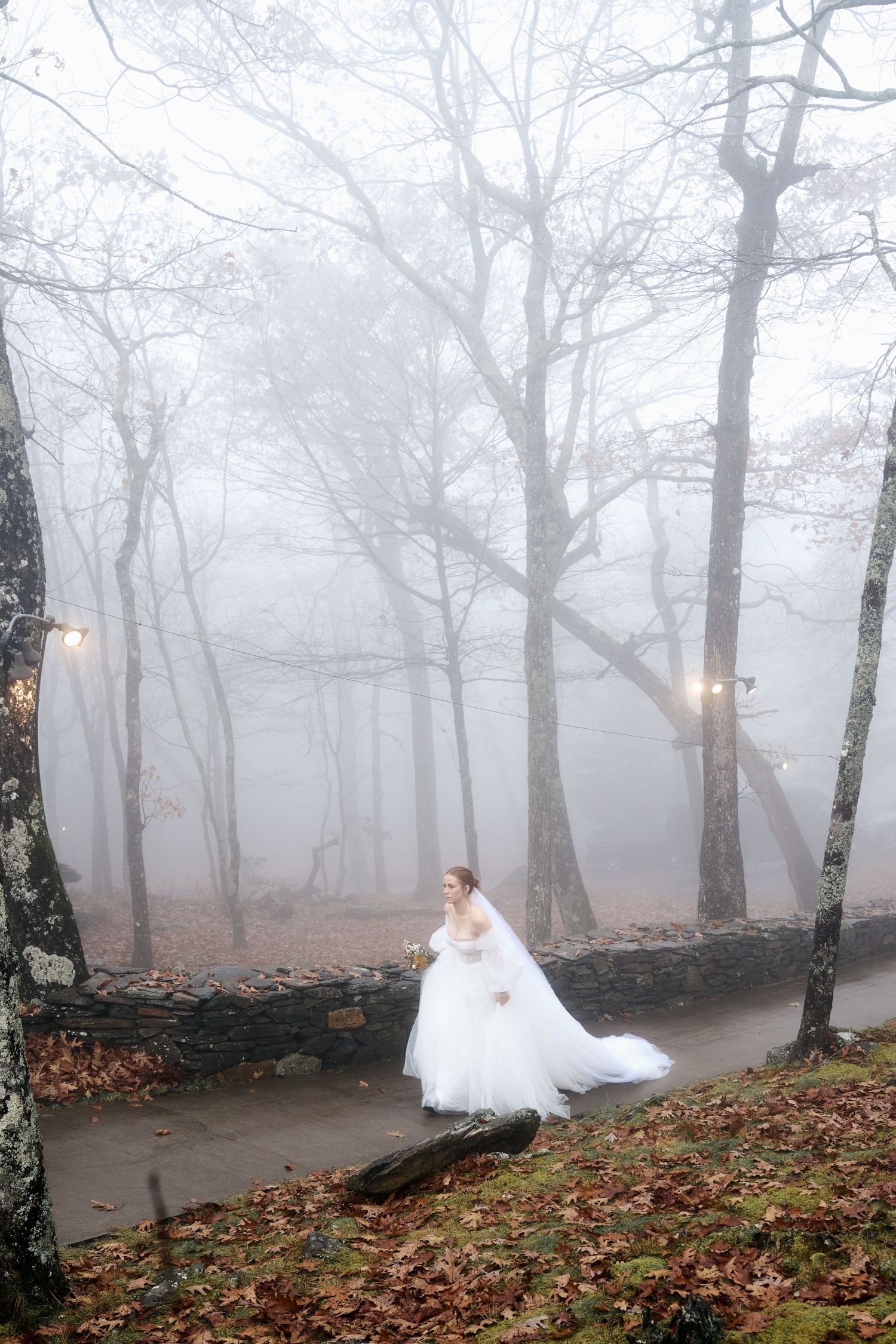 A bride in a flowing white gown walks through a misty forest with glowing lanterns.