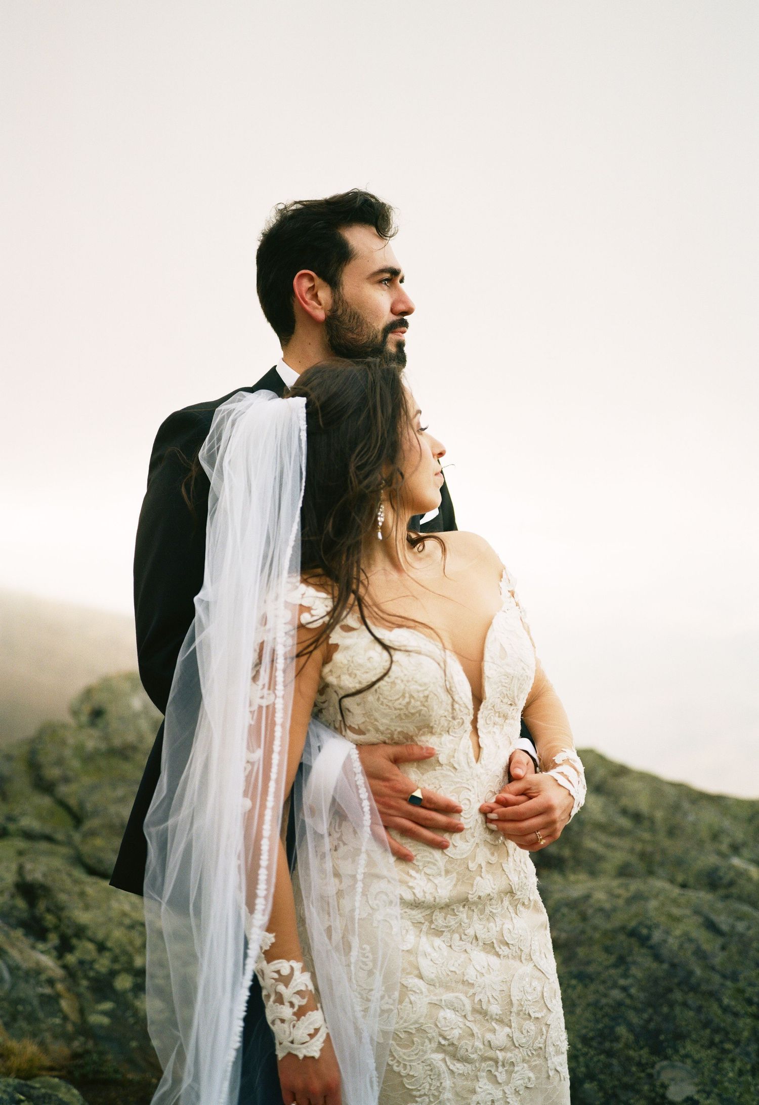 Romantic wedding couple embraces on a hillside at sunset in a lace gown and dark suit.