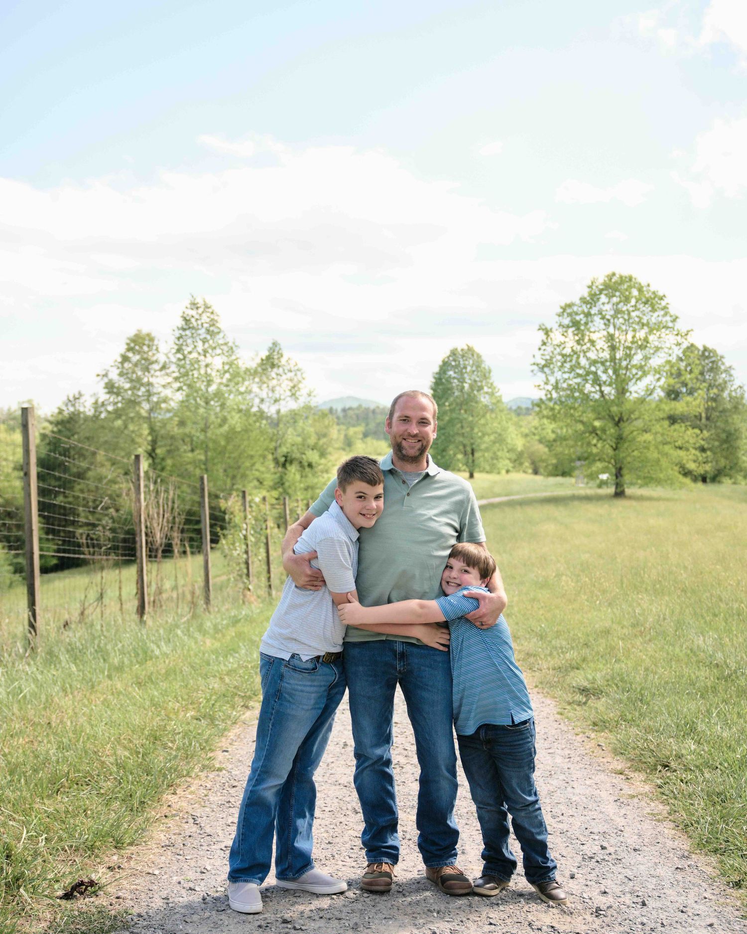 A man and two boys hug and smile on a gravel path surrounded by green fields and trees.