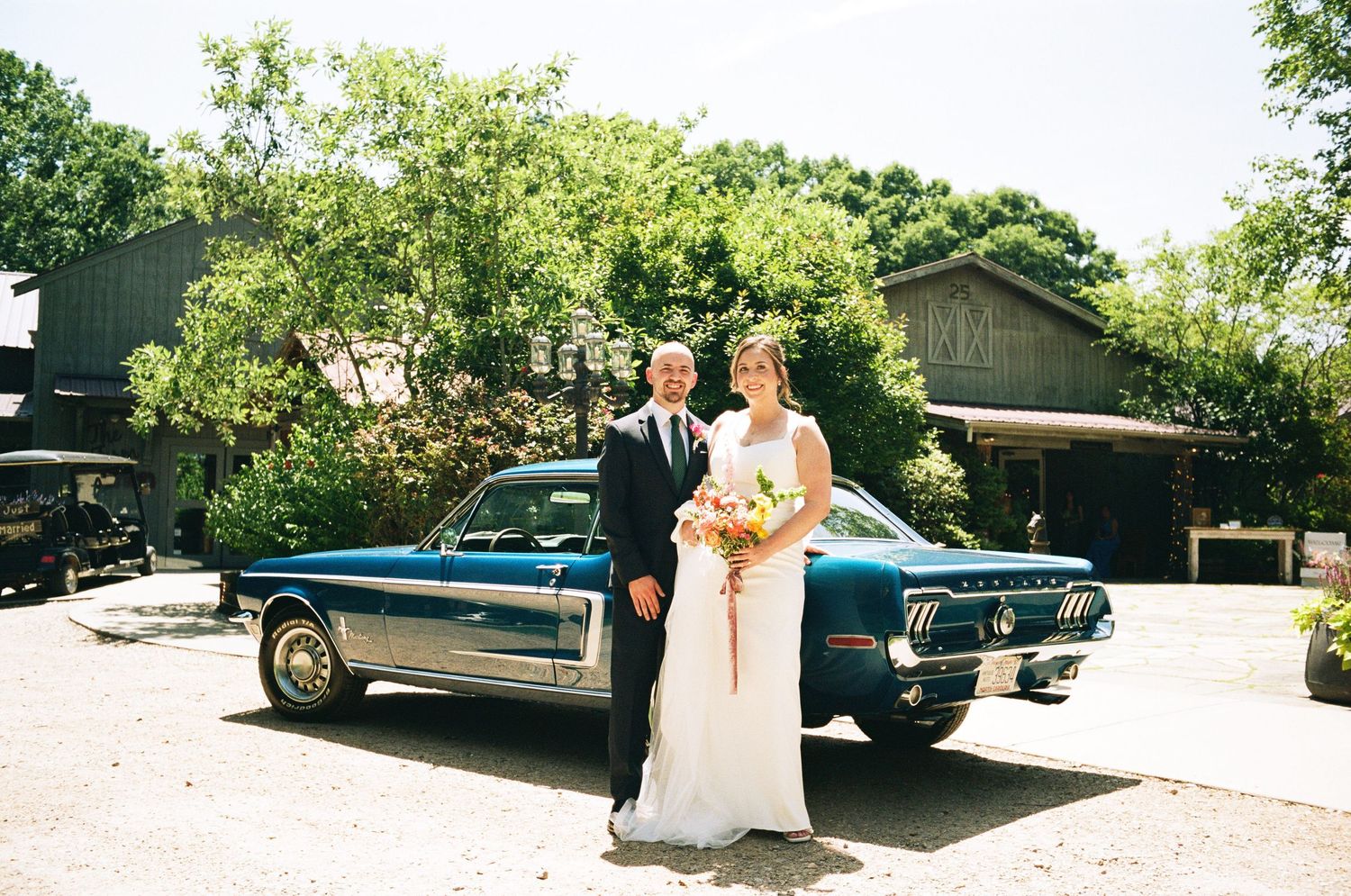 Bride and groom posing beside a classic teal Ford Mustang on their wedding day outdoors.