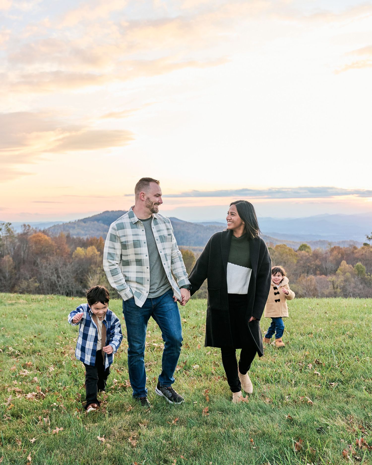 A family enjoys a playful moment together on a grassy hilltop with mountains and autumn trees in the background at sunset.