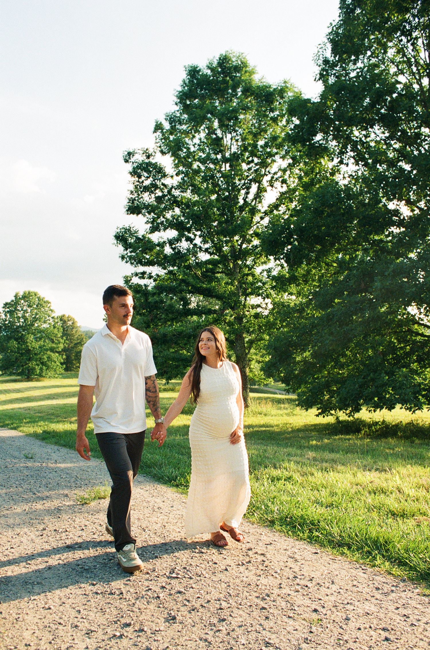 Pregnant couple holding hands walking on a sunny gravel path surrounded by lush green trees.