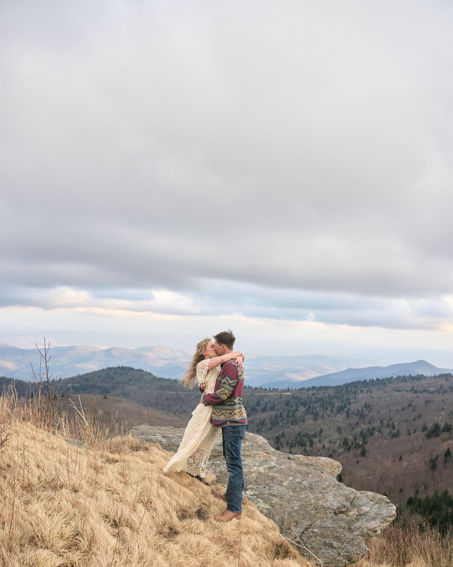 A couple embraces while standing on a mountaintop overlooking a scenic mountain landscape during cloudy weather.