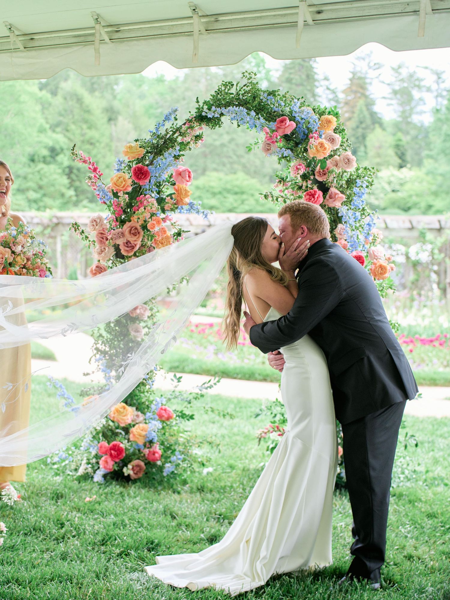 A romantic couple shares a tender kiss under a floral-decorated outdoor canopy on a grassy lawn during their wedding ceremony.