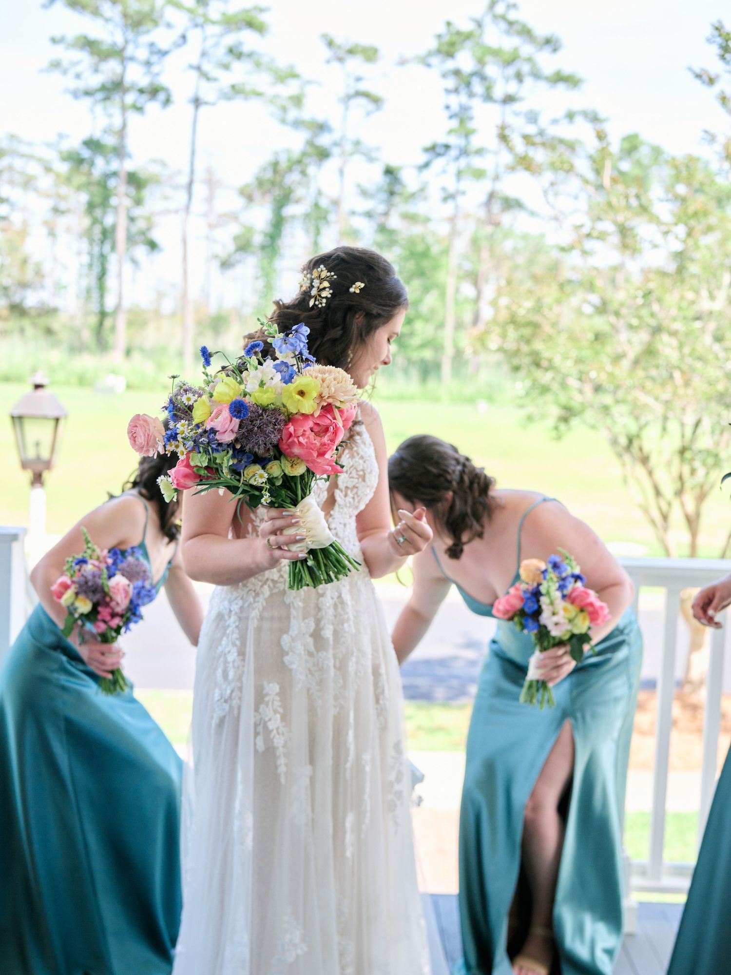 Wedding party in turquoise bridesmaid dresses holds colorful bouquets with pink and blue flowers outdoors by pine trees.