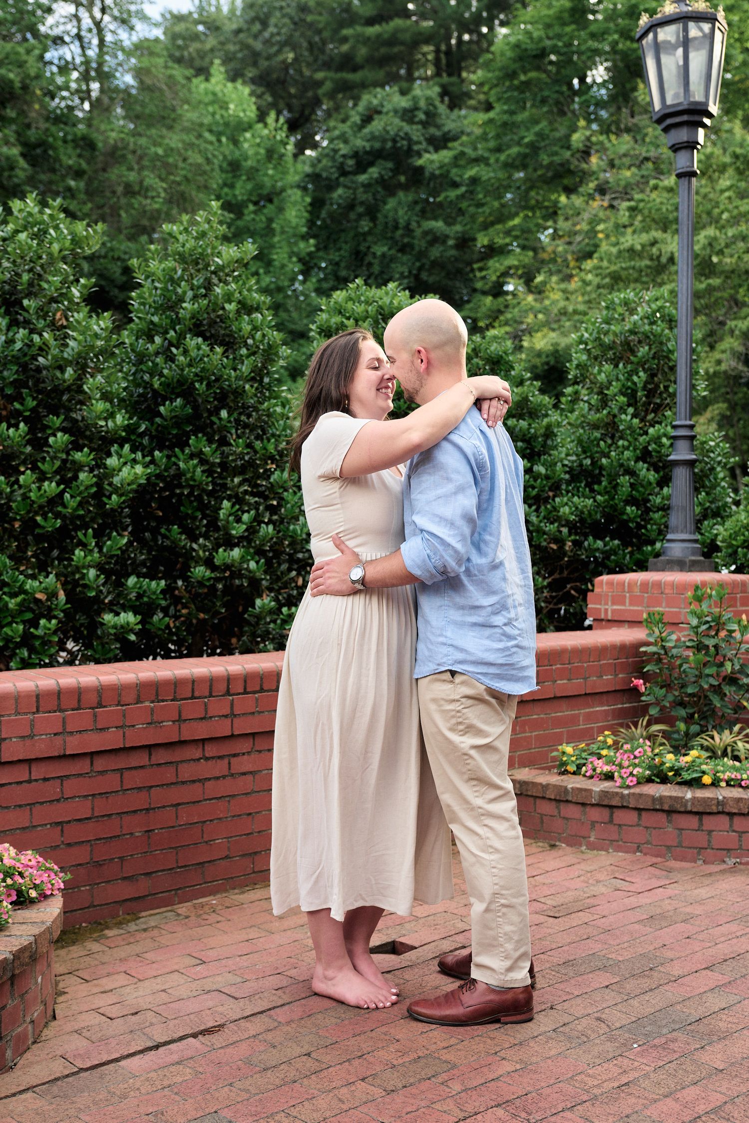 A couple embraces on a brick patio surrounded by gardens and a vintage lamppost.