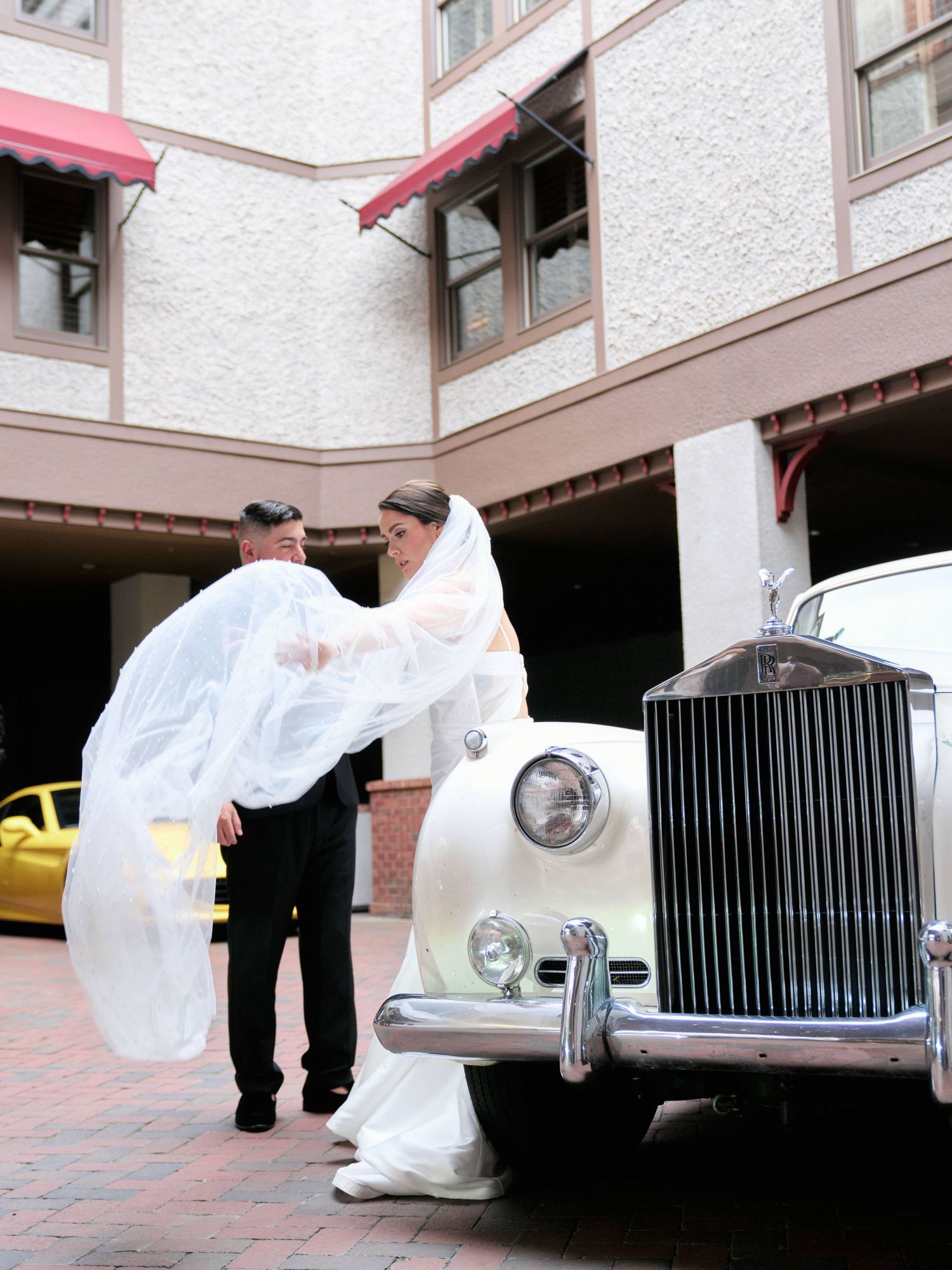 A classic white Rolls-Royce parked at an entrance with people in wedding attire standing beside it.