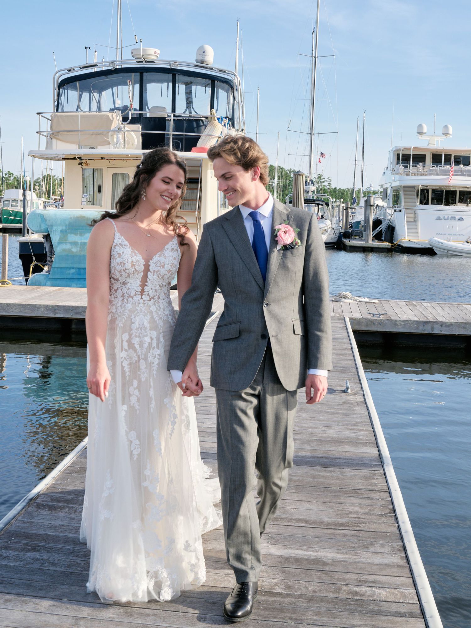A couple in wedding attire walk together on a wooden dock at a marina with boats in the background.