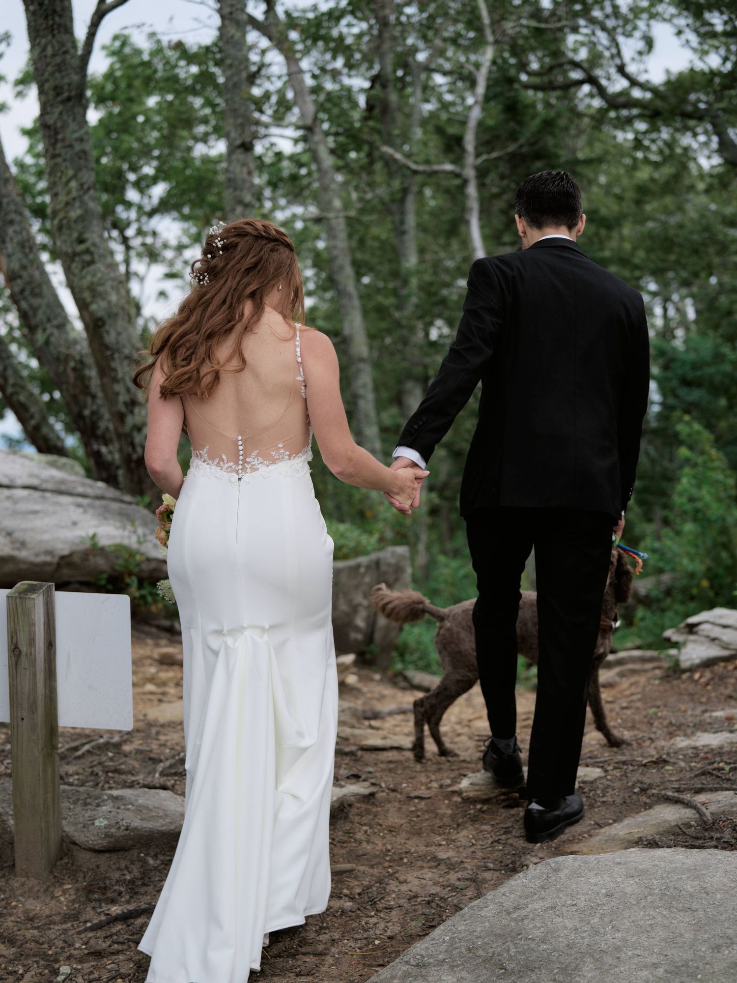 A couple in wedding attire walks together on a rocky woodland path during their outdoor ceremony.