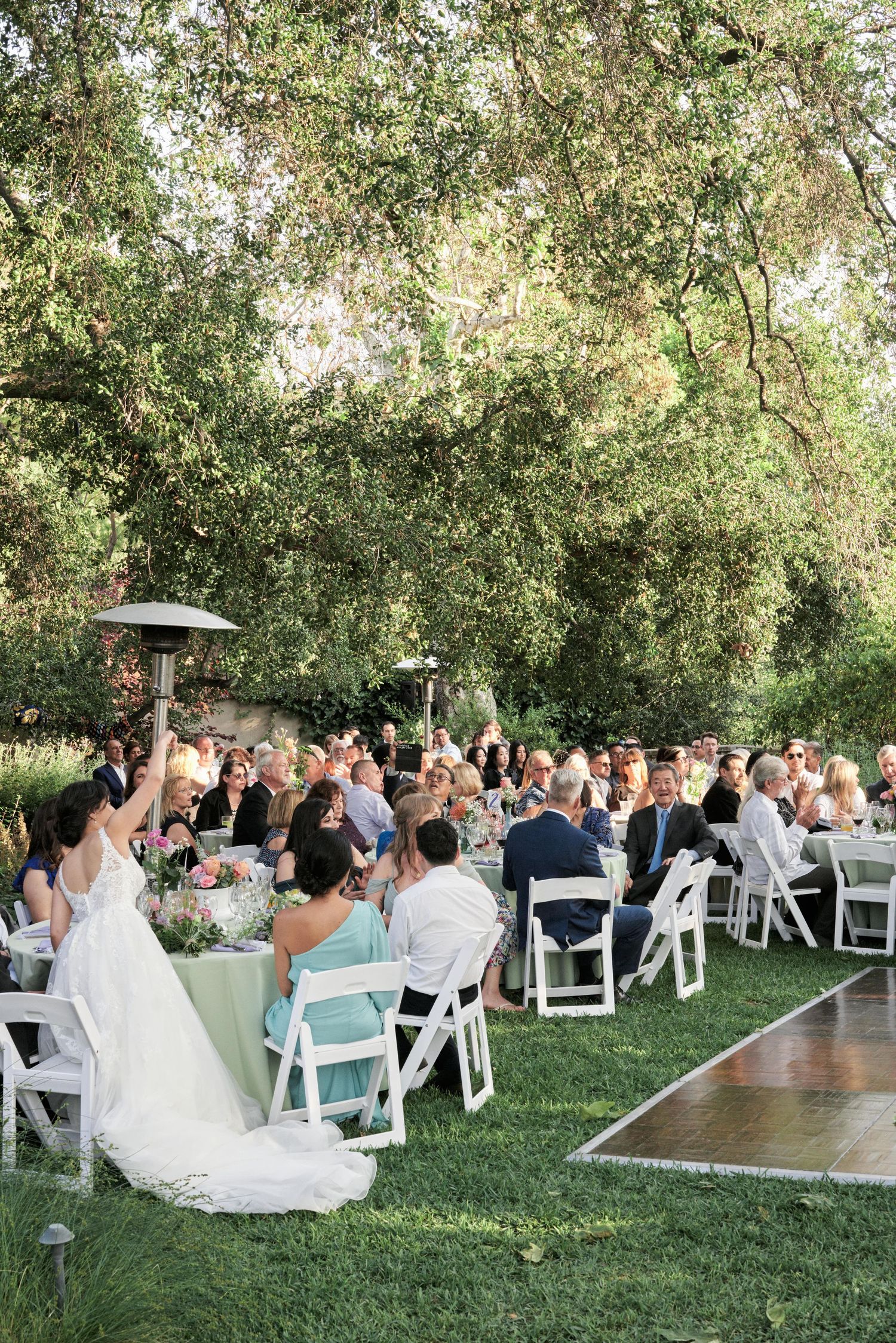 Outdoor wedding reception under trees with guests seated at white chairs and tables on a grassy lawn.